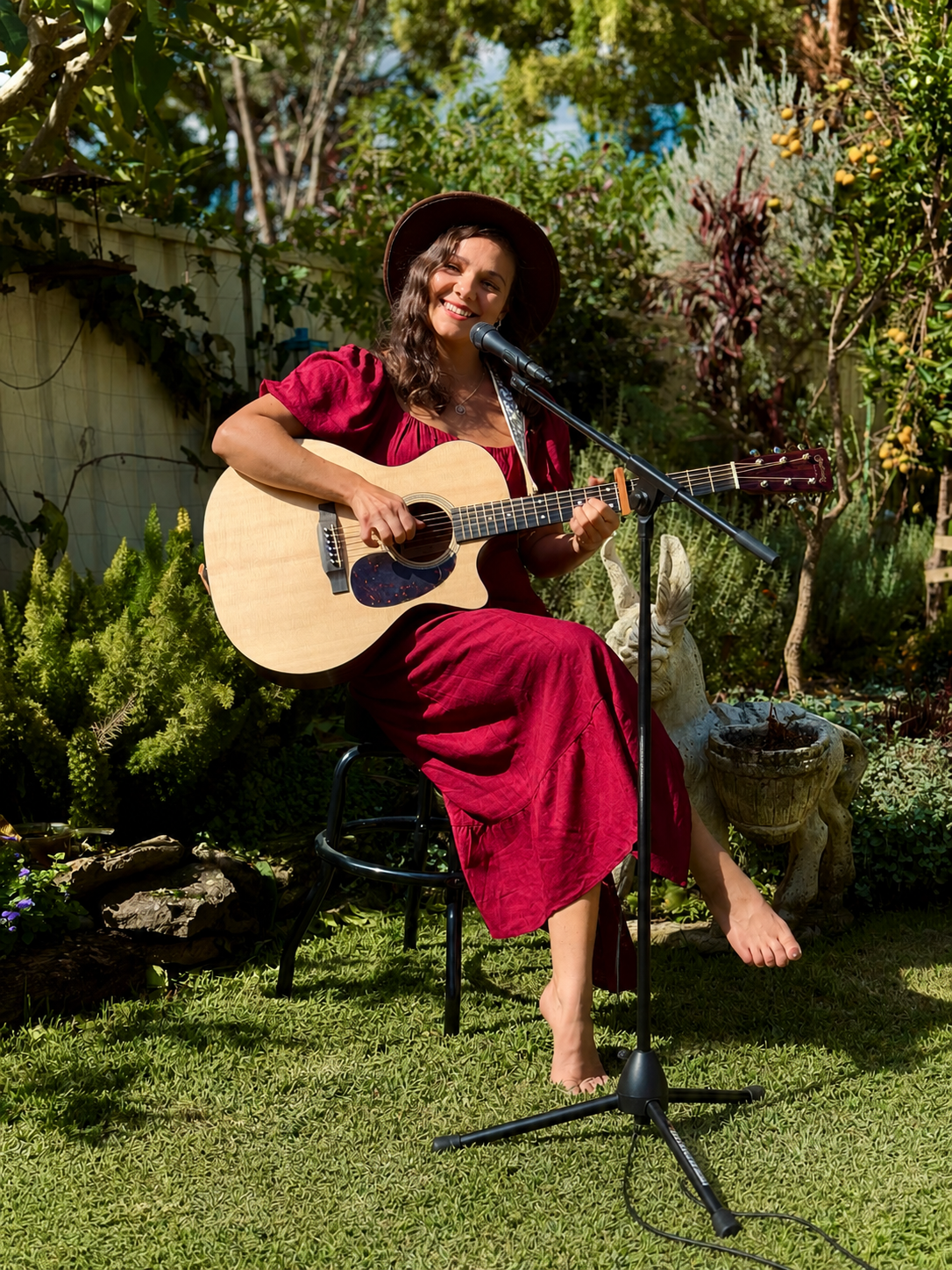 A woman in a red dress and wide-brimmed hat playing an acoustic guitar and singing into a microphone in a lush garden.