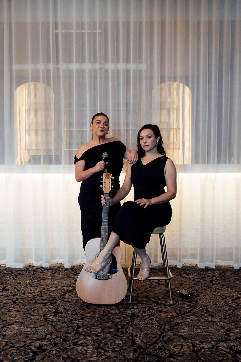 Two women in elegant black dresses are posing indoors against sheer curtains. One woman holds a microphone and a guitar, while the other sits on a stool with one leg crossed over the other.