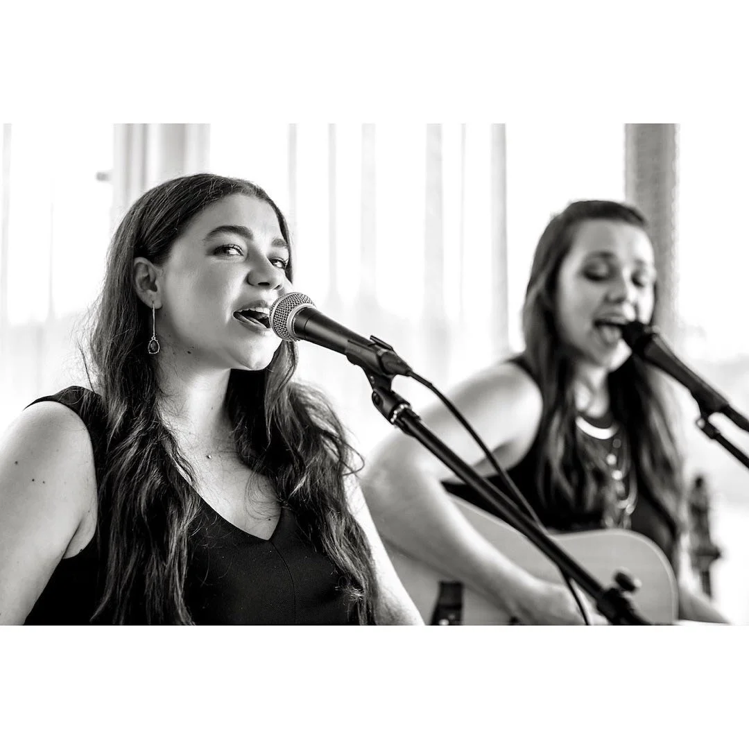 Two women singing into microphones; one plays an acoustic guitar; black and white photo.