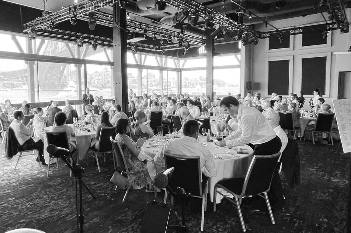 A formal gathering or banquet in a large, well-lit room with floor-to-ceiling windows, round tables with people eating and drinking, a speaker at a podium, and a stage with lighting equipment.