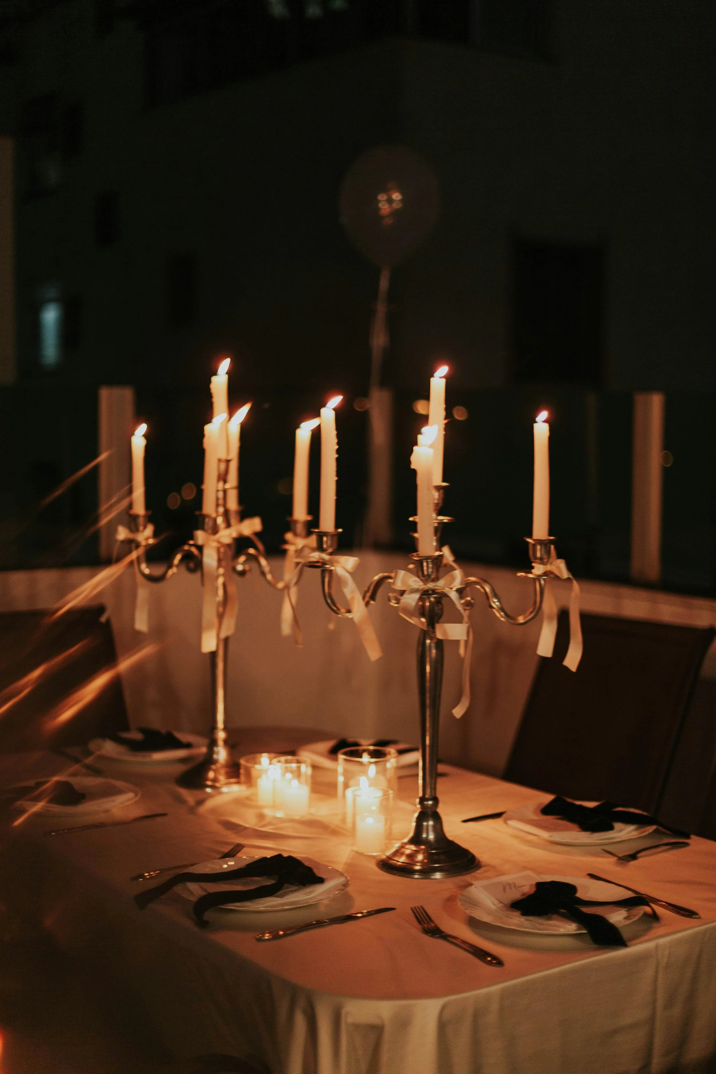 A dinner table decorated for a celebration with a silver candelabra holding lit candles, surrounded by plates, silverware, black napkins, and candles in small glass holders, set in a dimly lit room.