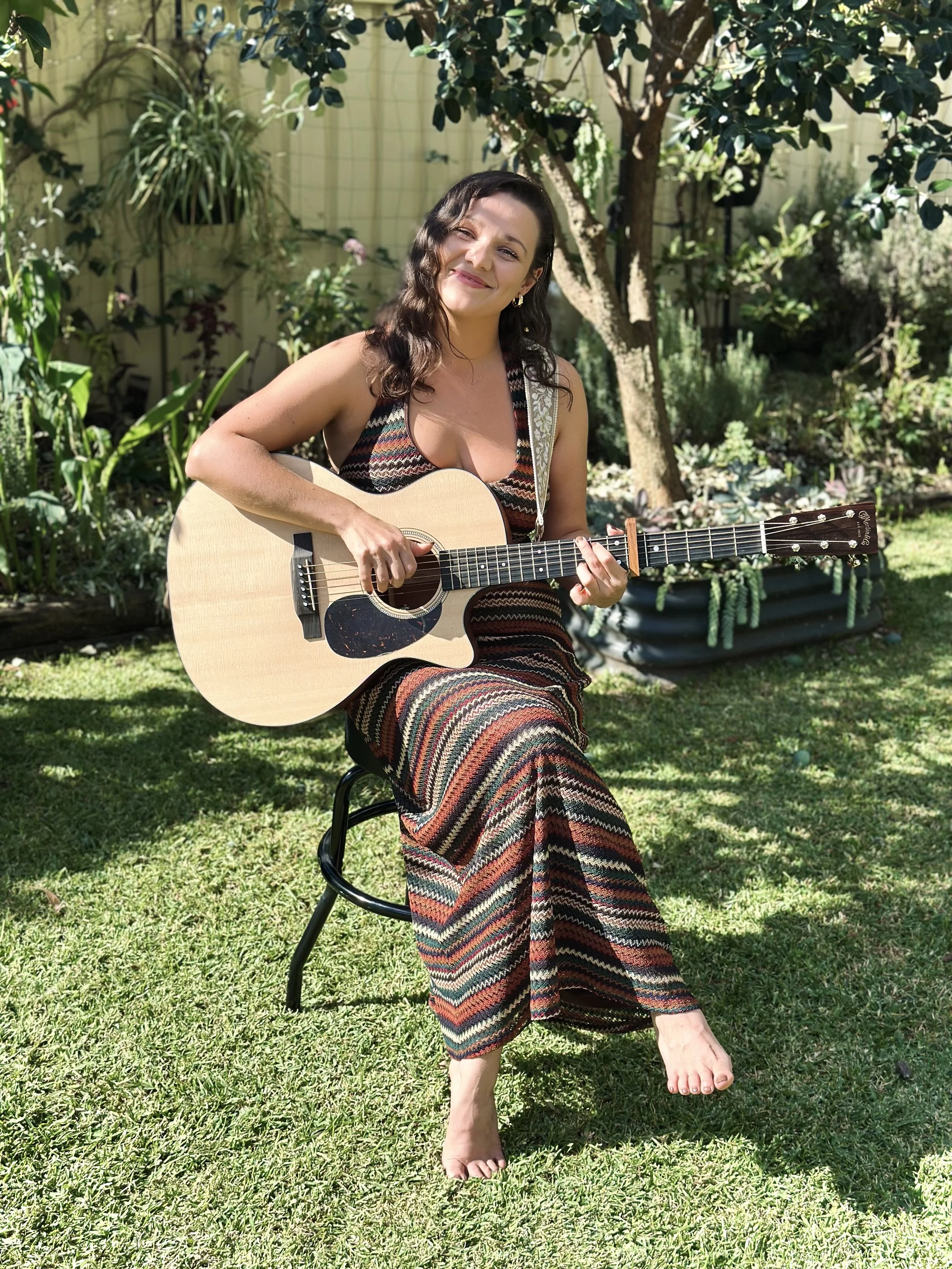A woman with dark wavy hair, smiling, sitting outdoors on a black chair, playing an acoustic guitar in a garden with green plants and trees, wearing a long patterned dress.