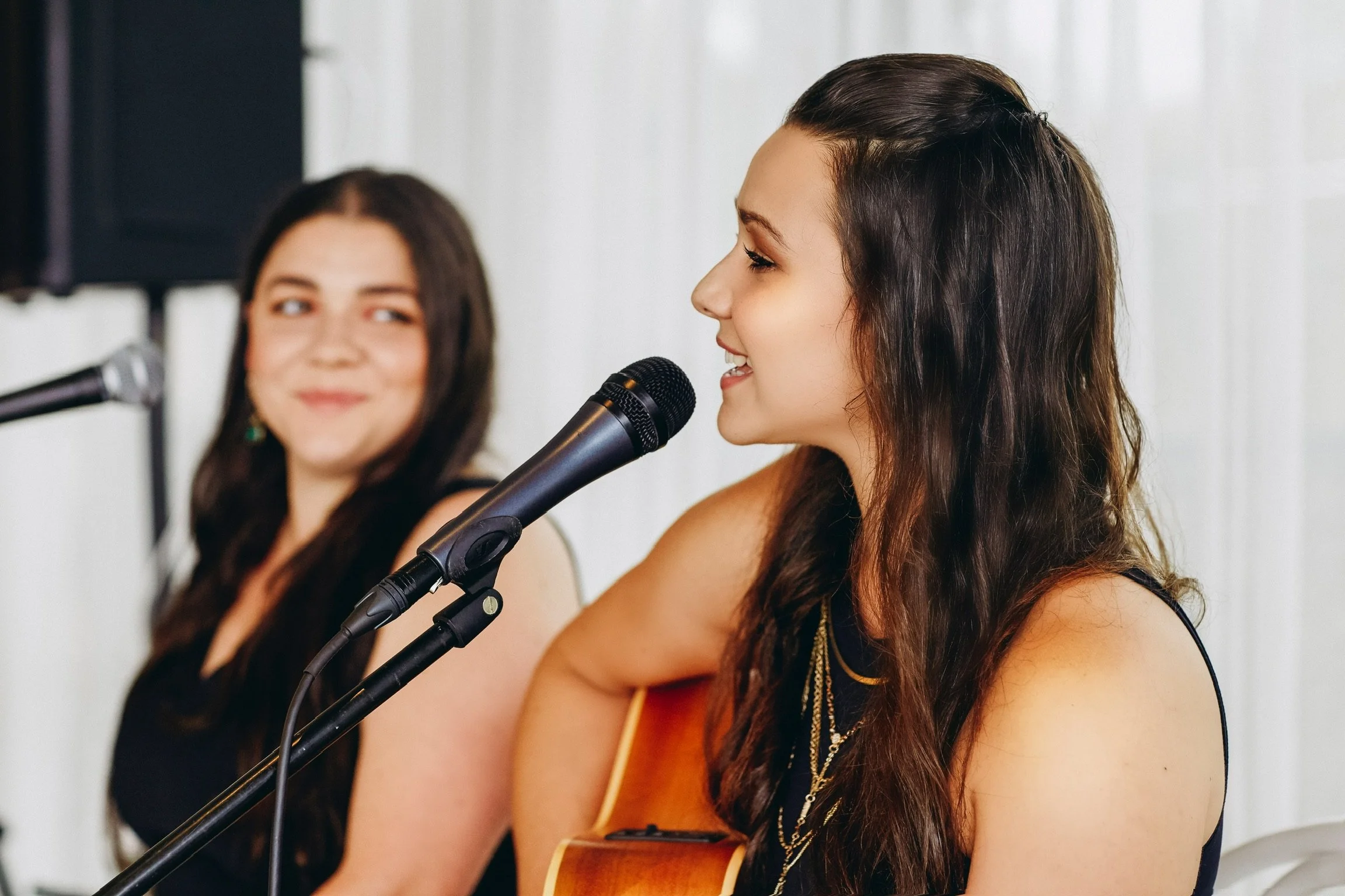 Two women are sitting at a table with microphones, one woman is singing or speaking into a microphone while wearing a sleeveless top and a necklace, and the other woman is smiling and looking at her.