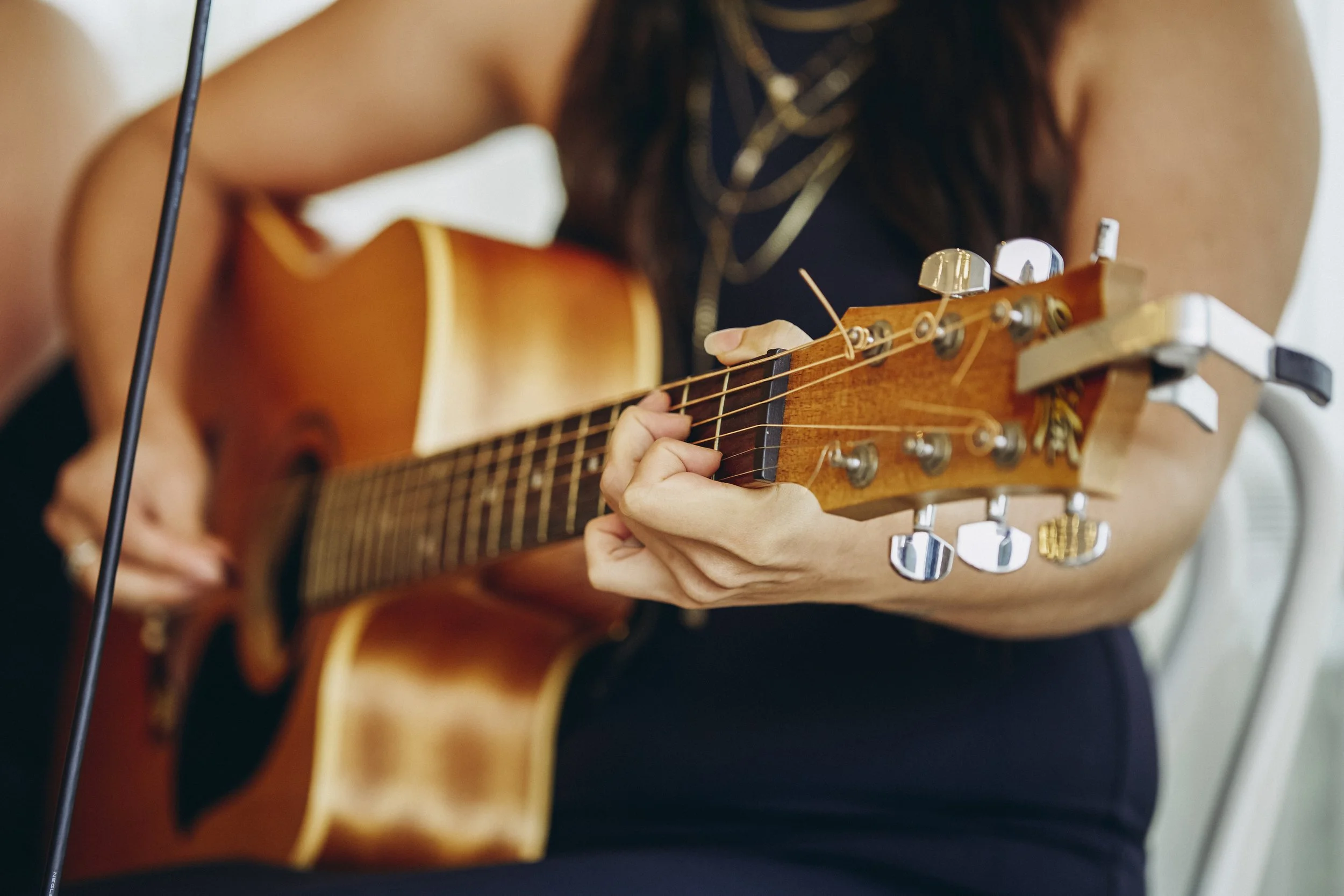 A person playing an acoustic guitar, with focus on their hand on the fretboard and the headstock of the guitar, wearing a dark top and multiple layered necklaces.