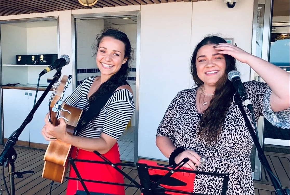 Two women smiling, one holding a guitar, on a balcony with microphones and music stand, with a door and exterior view in the background.