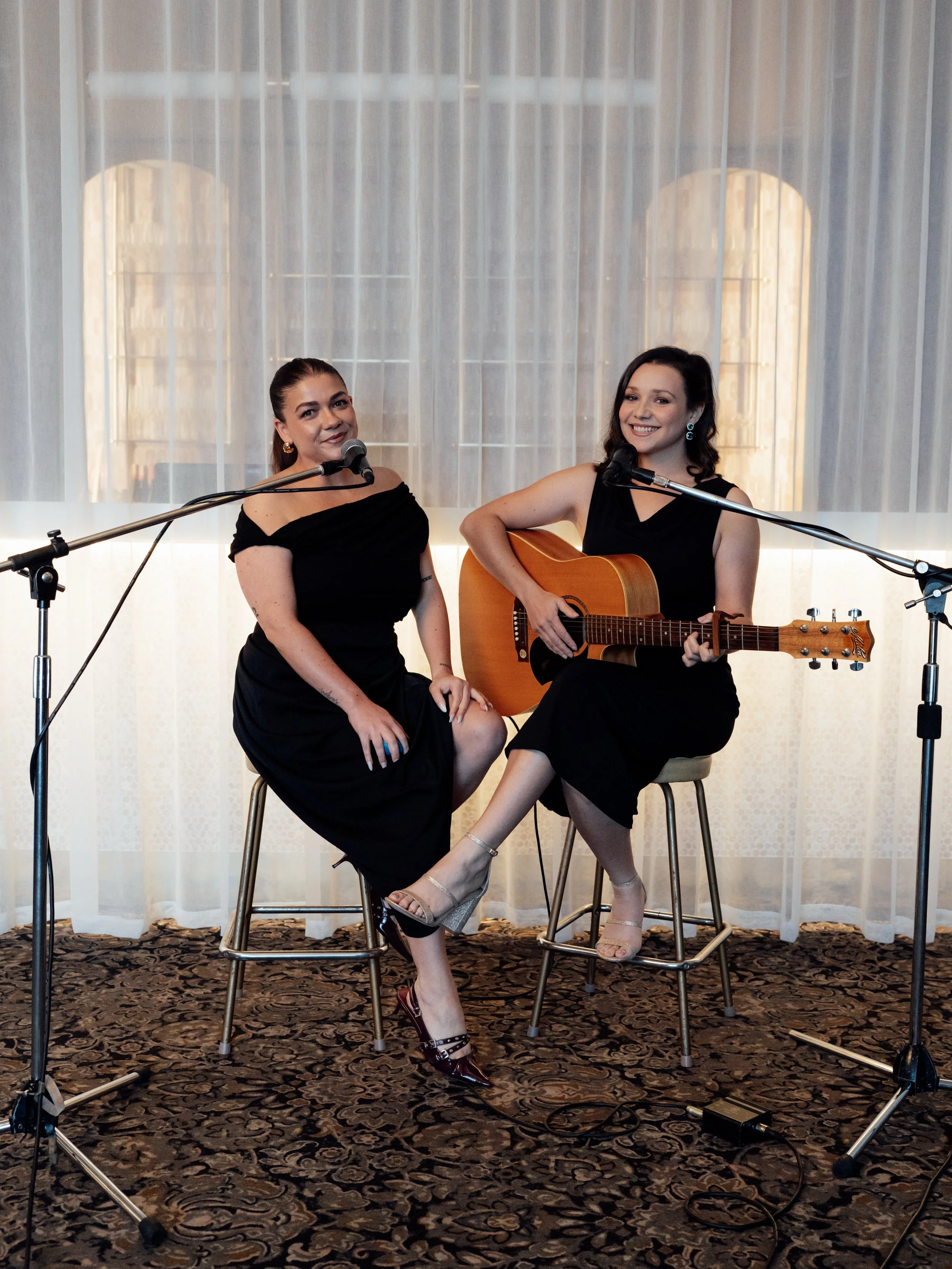 Two women wearing black dresses performing with microphones and a guitar in a room with a patterned rug and sheer curtains.