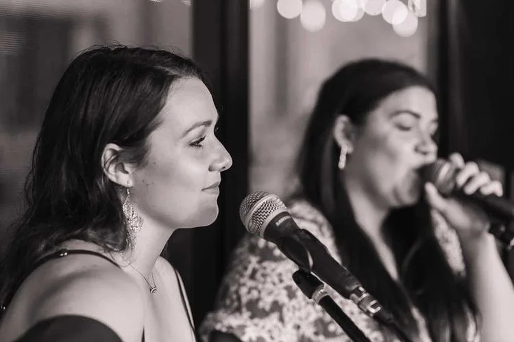Two women singing into microphones at a social gathering, one in focus and the other slightly blurred in the background.