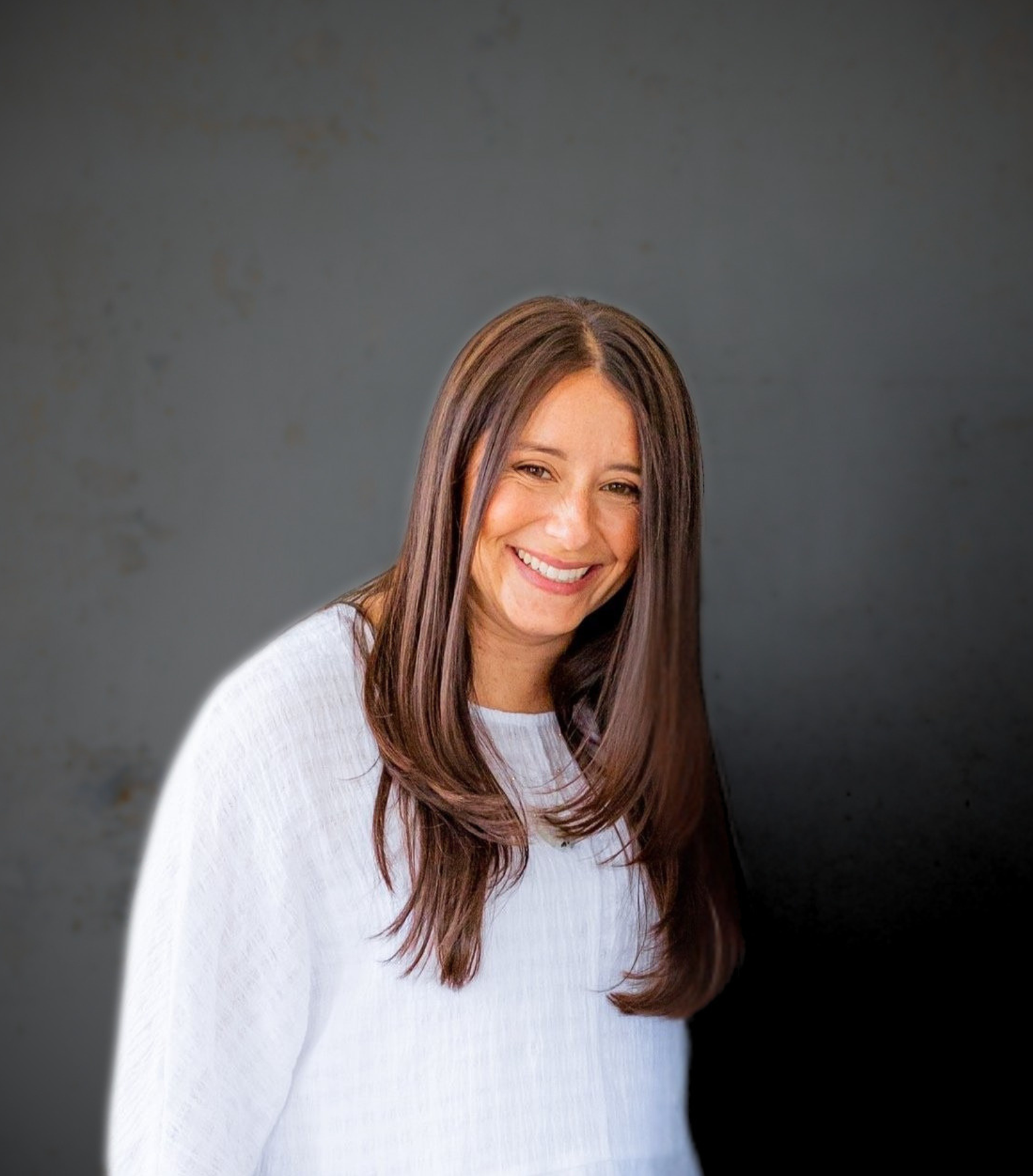A woman with long brown hair smiling, wearing a white top, against a dark gray background.