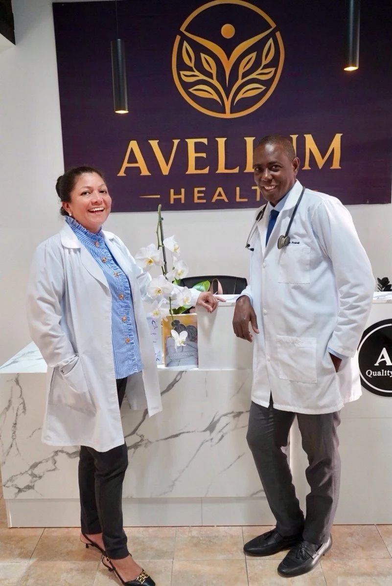 Two medical professionals in white coats stand in front of a reception desk at Avellum Health, smiling at the camera. One is a woman with dark hair, and the other is a man with a stethoscope. The background features a dark purple wall with a gold logo and the words 'AVELLUM HEALTH'.