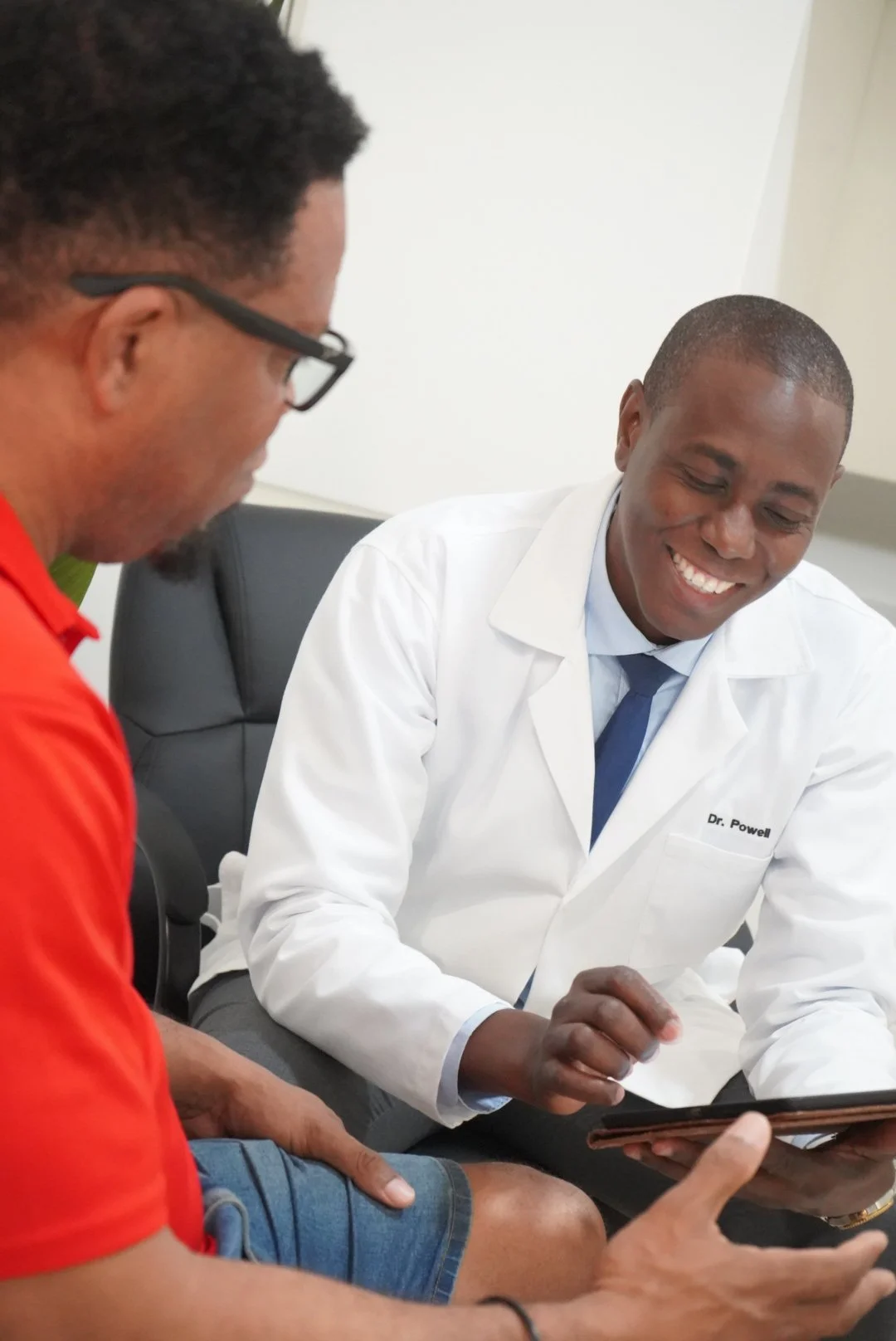 Two men sitting and looking at a tablet, one wearing a white coat with 'Dr. Powell' on it, and the other in a red shirt with glasses, smiling.