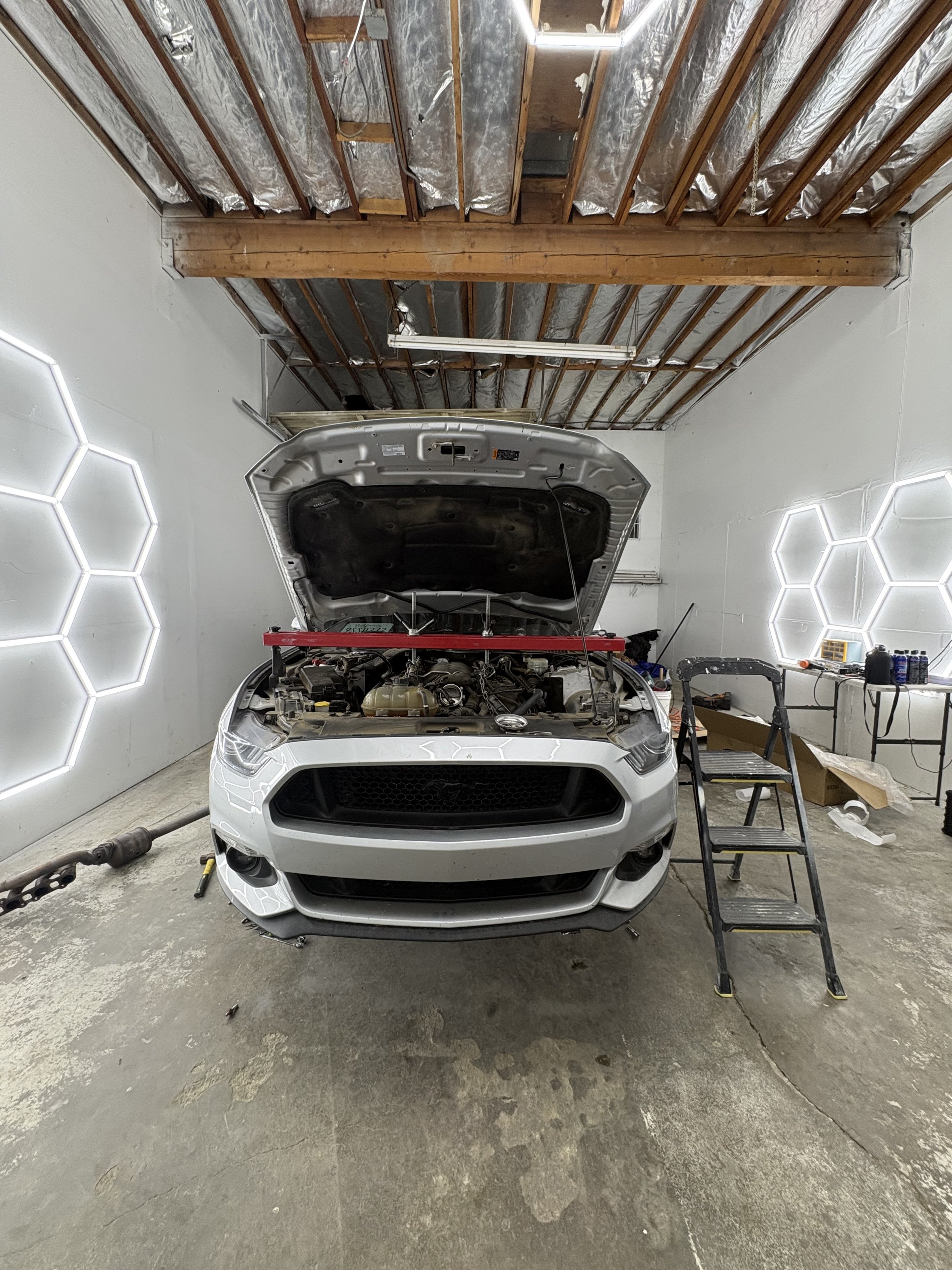 White car with open hood in a garage or workshop, with tools and a step ladder nearby, and illuminated hexagonal wall lights.