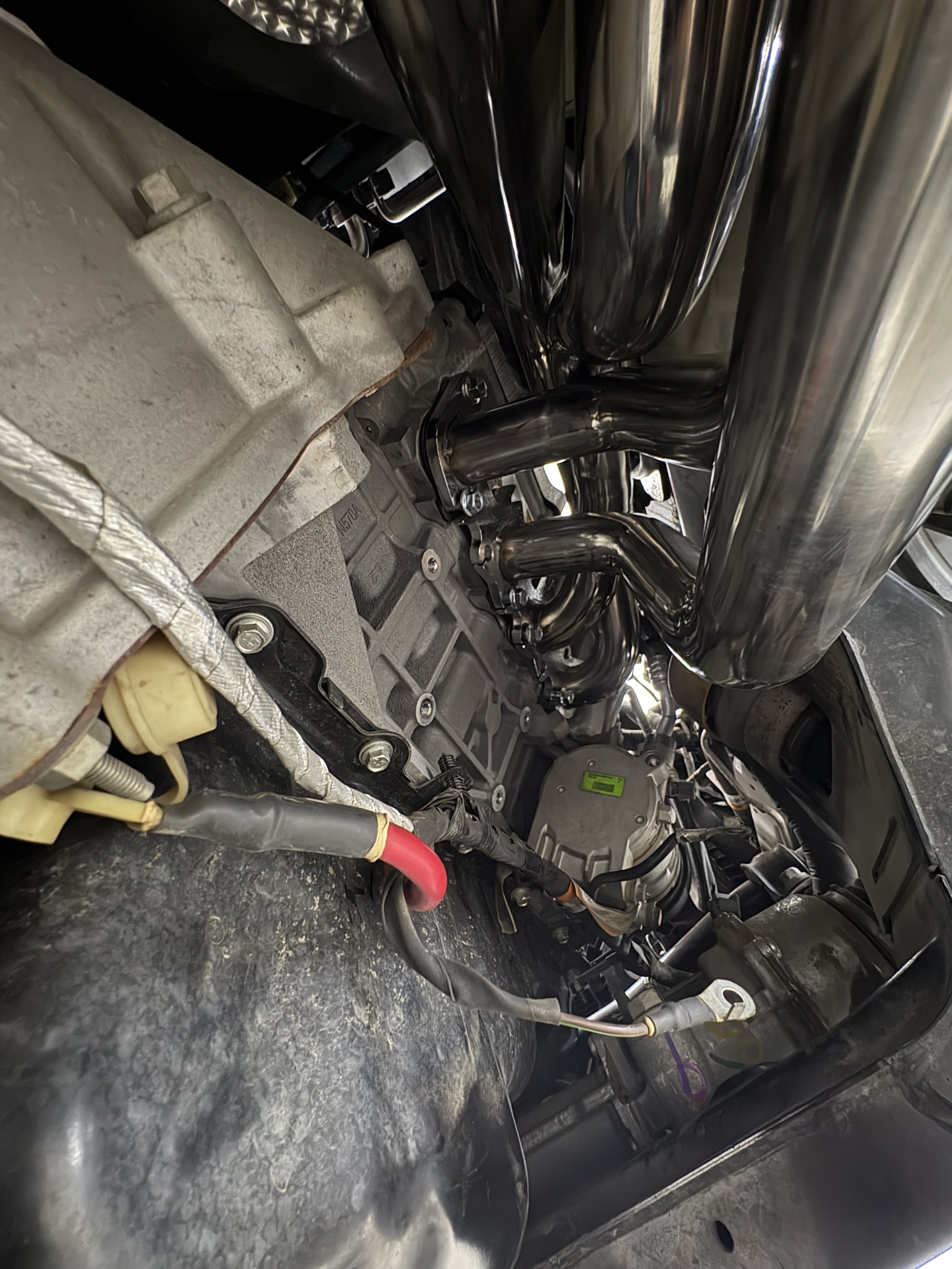 Close-up of a vehicle's engine bay showing black exhaust headers, the transmission, and various hoses and wires.
