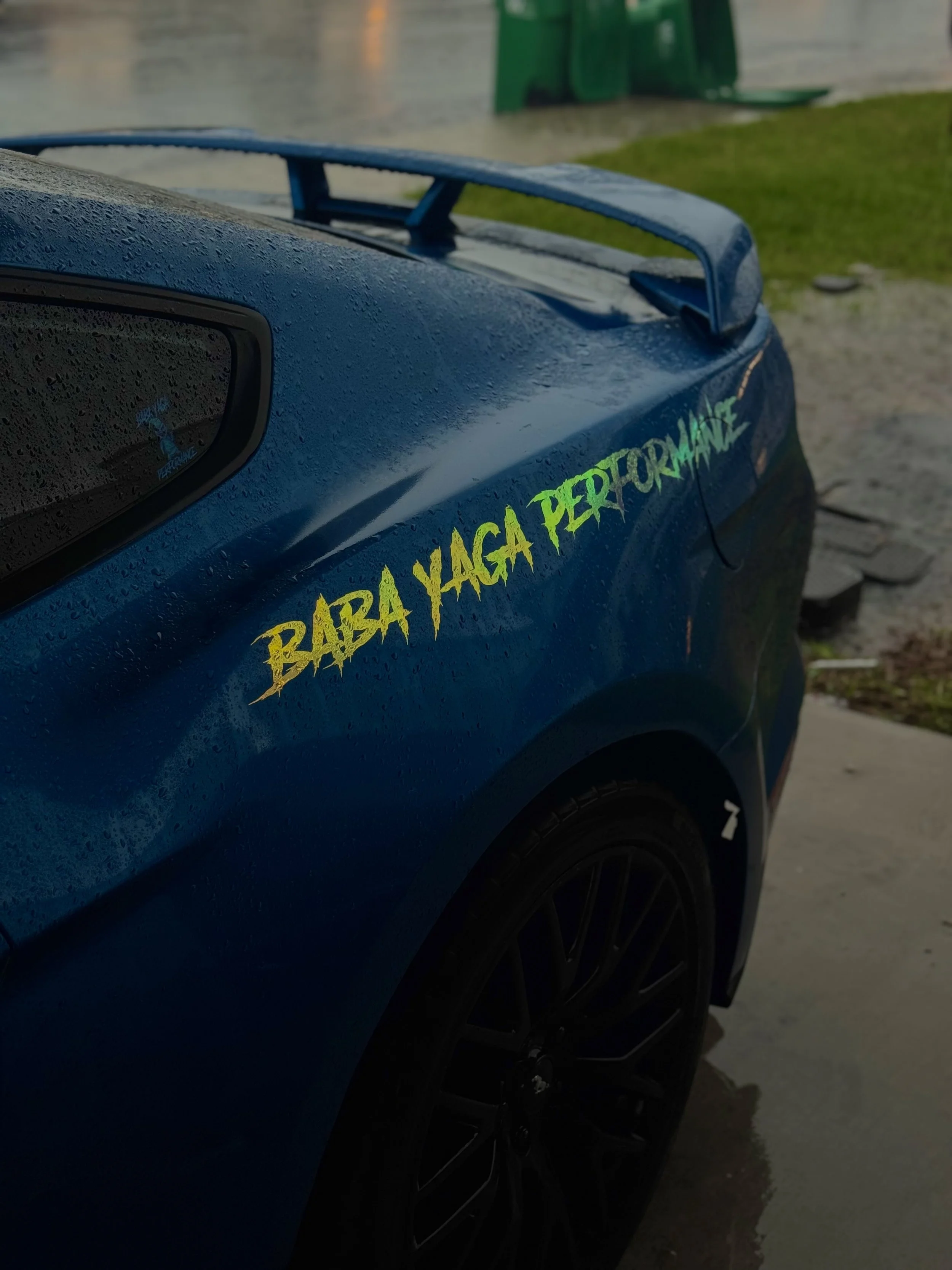 Close-up of a blue car with a yellow decal that reads 'BABY YAGA PERFORMANCE' on the side, with water droplets on the surface, wet pavement, and green structures in the background.