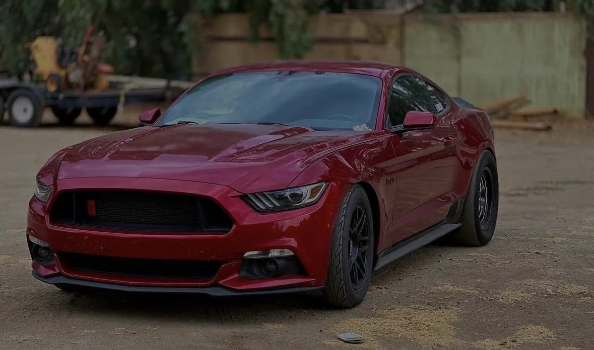 Red sports car parked on dirt ground with a wooden fence and trees in the background.