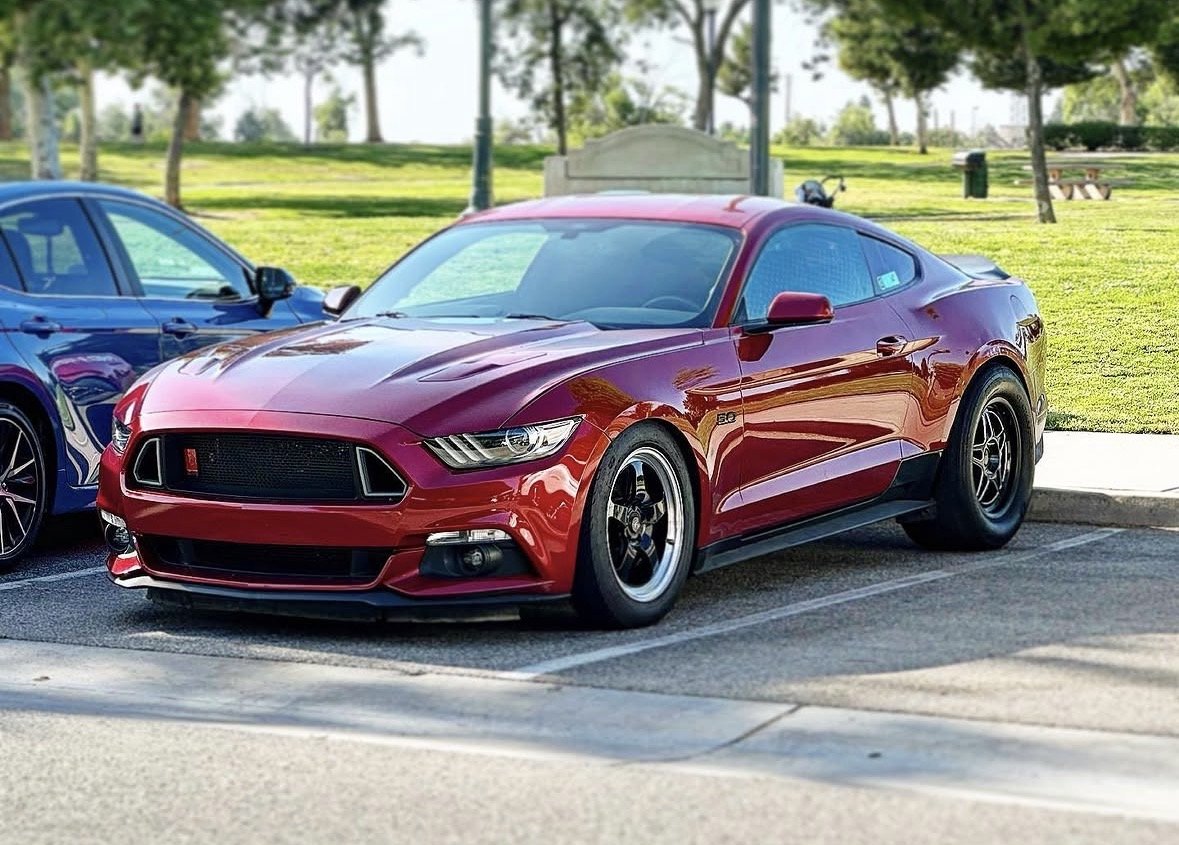 Red Ford Mustang parked in a parking lot near a grassy park with trees.
