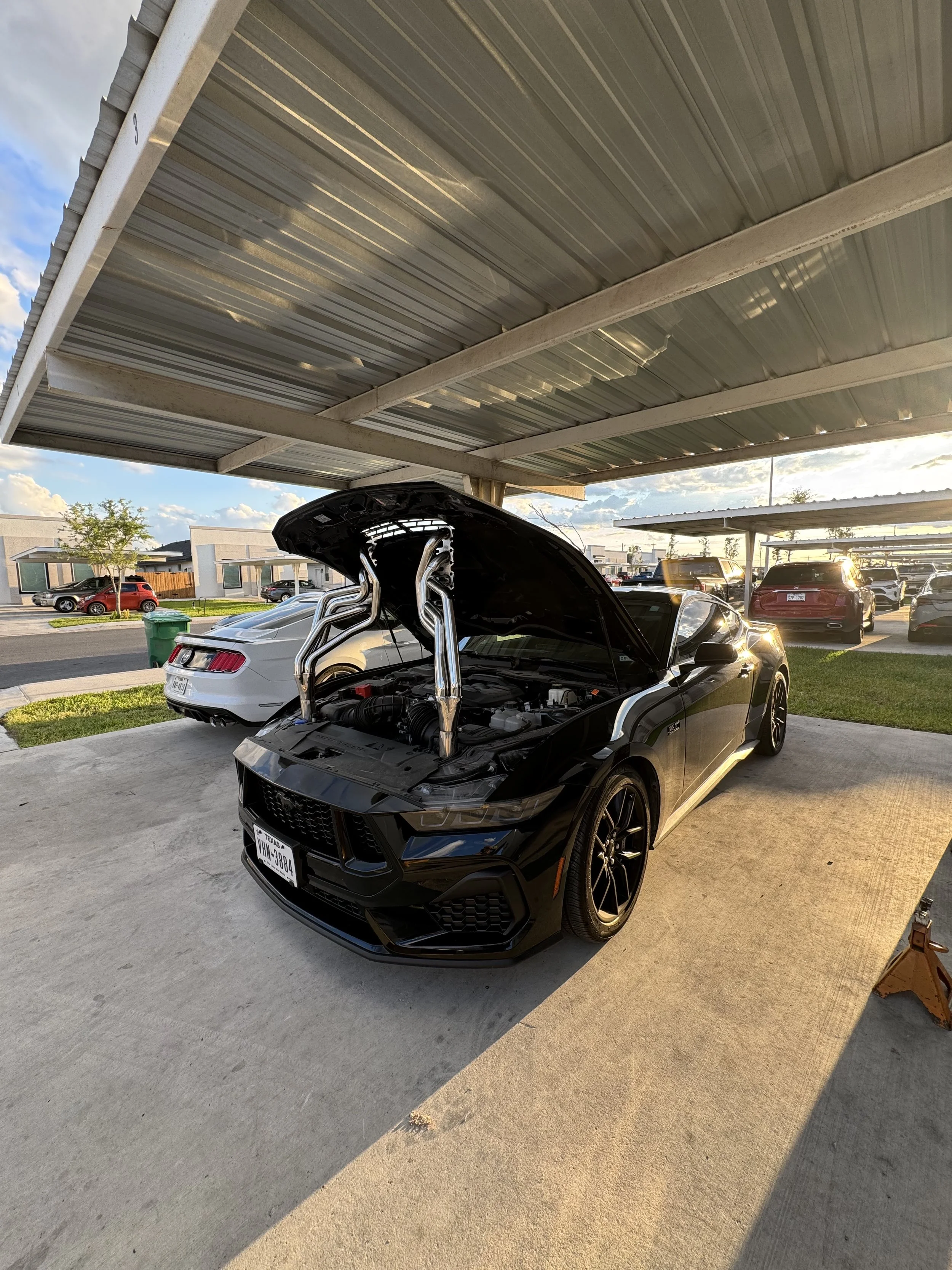 Black sports car with hood open under a metal carport, parked next to a white car, with other vehicles in the background