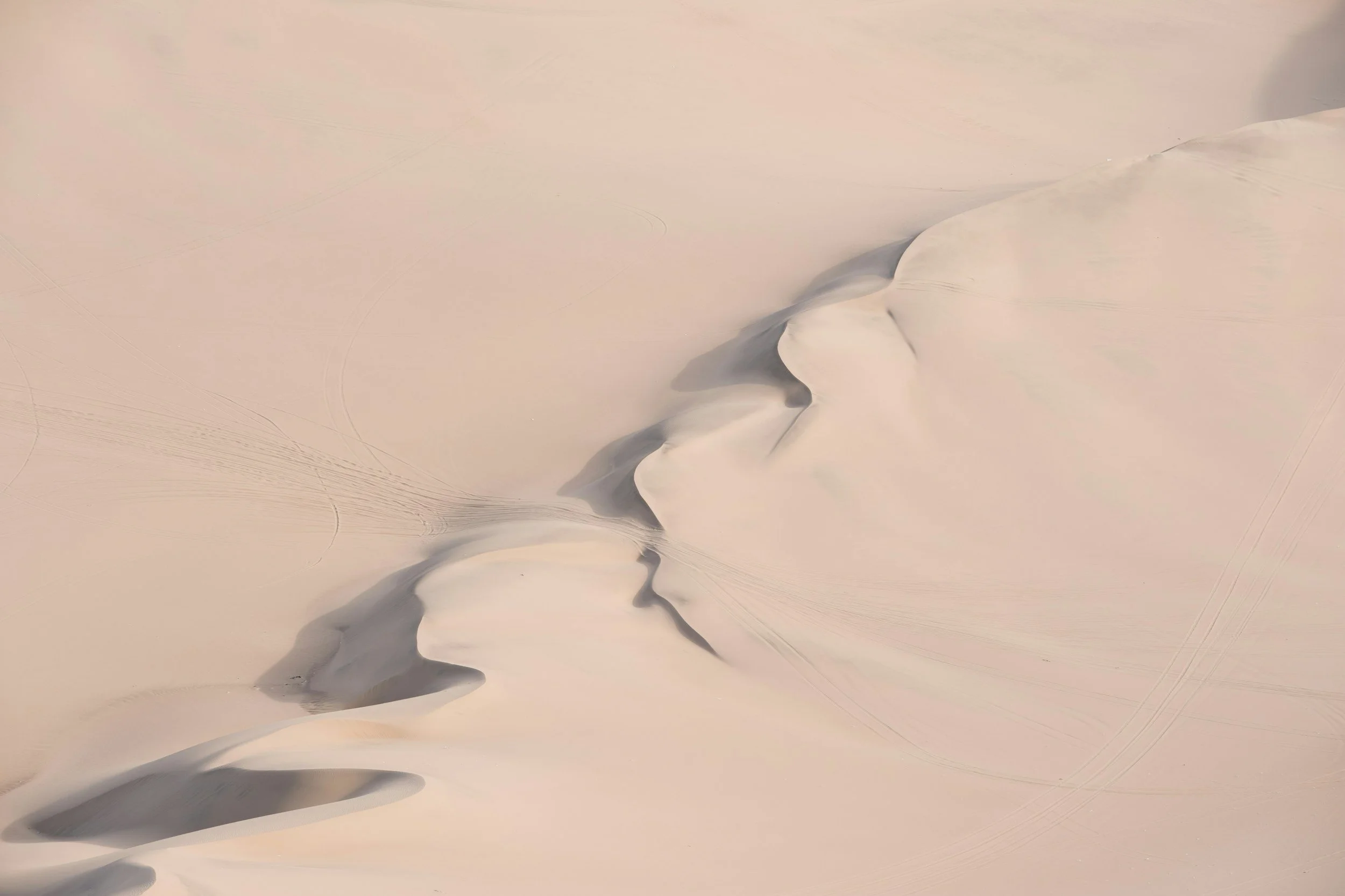 Aerial view of a vast white desert with rippled sand dunes and some dark streaks in the sand.