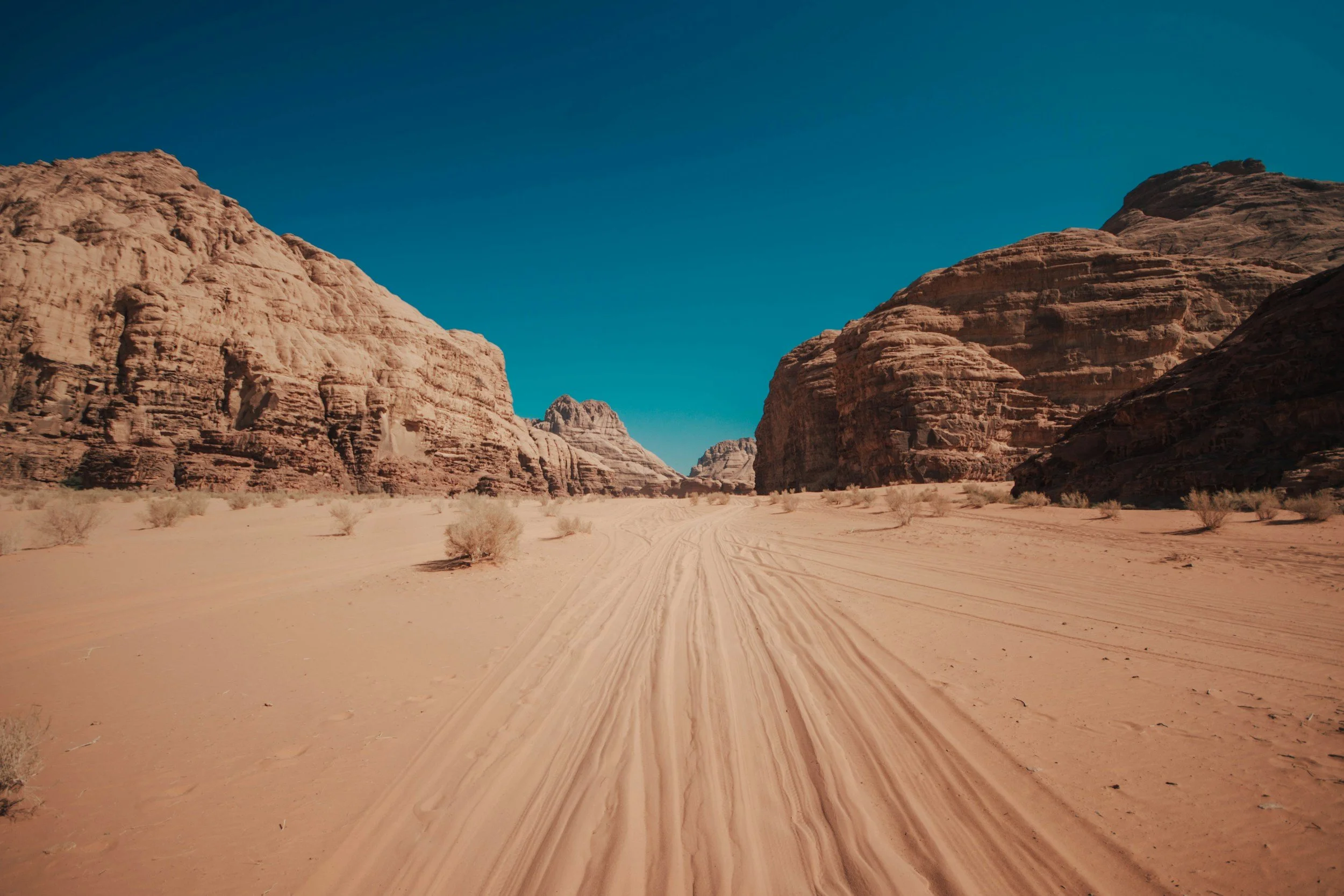 A desert landscape with large rock formations and a sandy, flat terrain with tire tracks, under a clear blue sky.