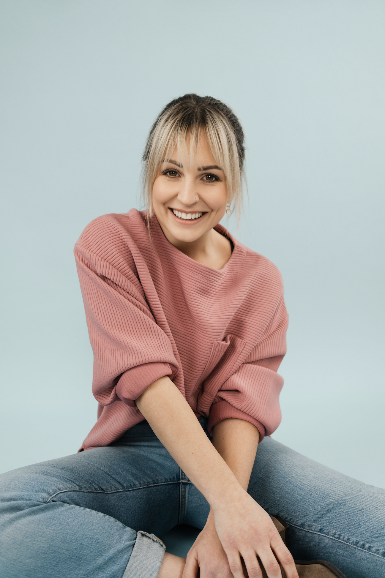 A woman with blonde hair in a ponytail, wearing a pink ribbed sweater with rolled-up sleeves, sitting on the floor against a light blue background and smiling at the camera.