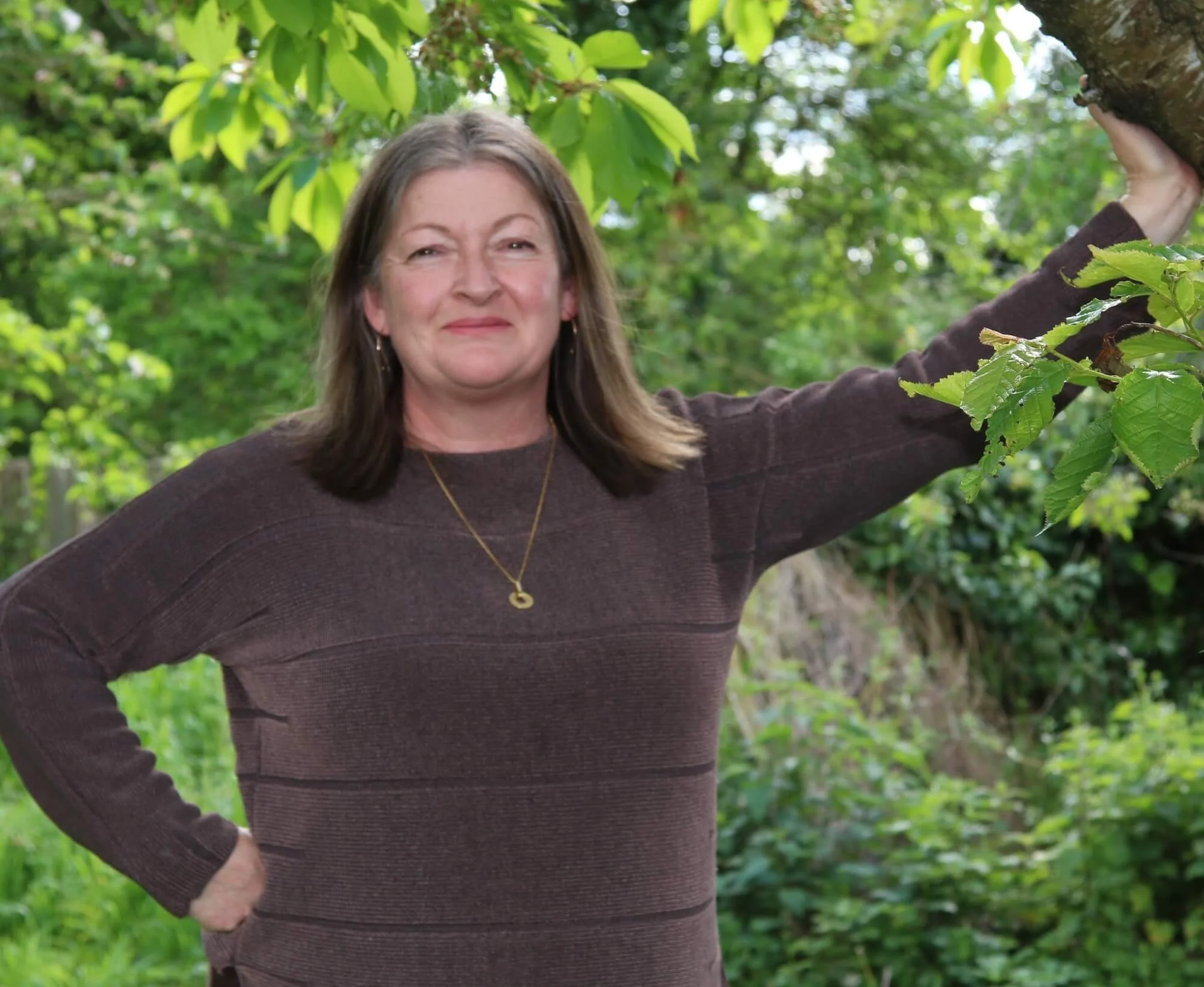 A woman with brown hair and a dark brown jumper standing outdoors in a lush green area, smiling with her hand on a tree branch