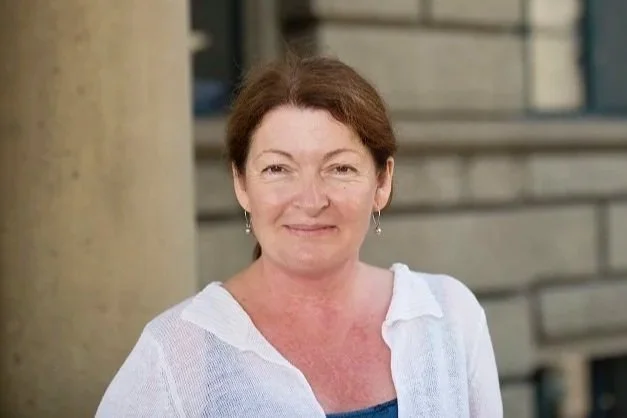 A woman with short brown hair, wearing a white cardigan over a blue top, standing outdoors in front of a building.