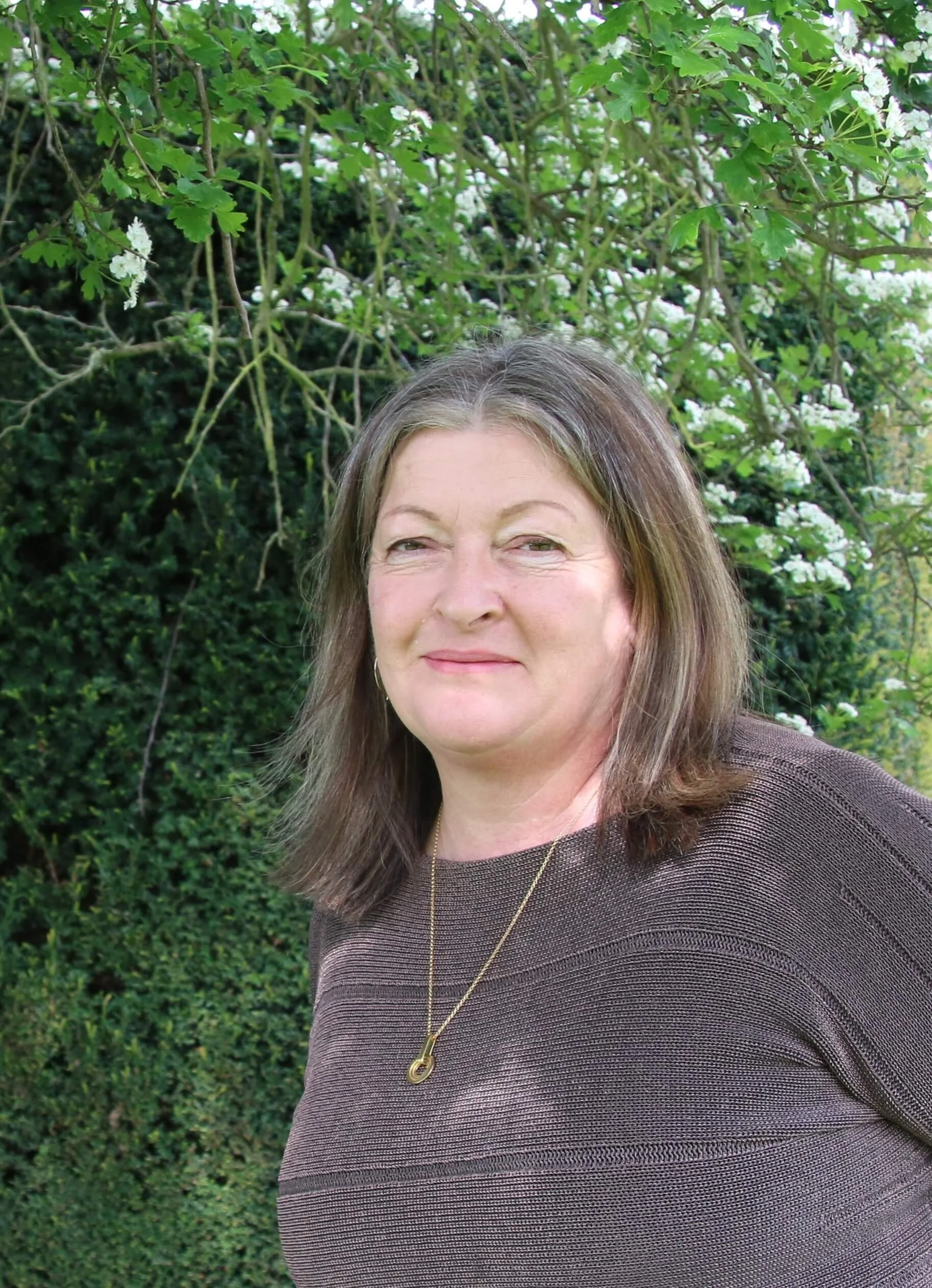 A woman with brown hair and a slight smile is standing outdoors in front of bushy greenery and white flowers.