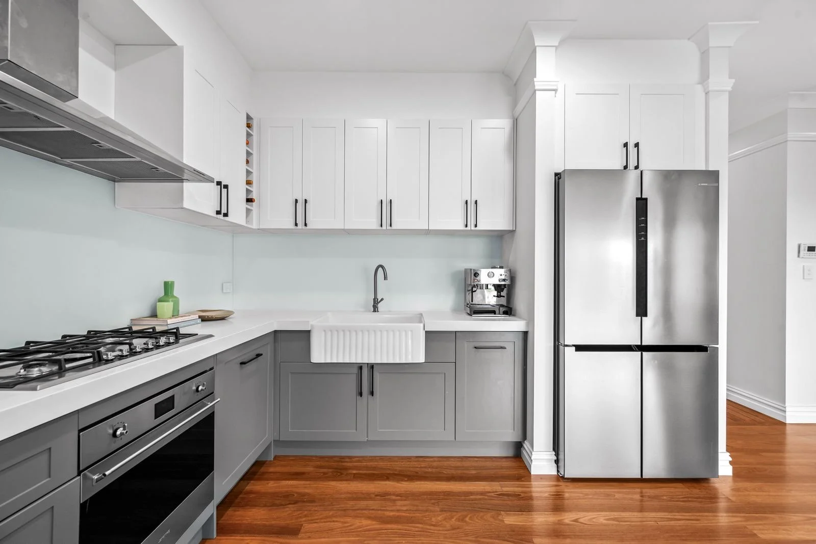 Modern kitchen with white upper cabinets, gray lower cabinets, stainless steel refrigerator, built-in oven, coffee machine, and a farmhouse sink on a white countertop.