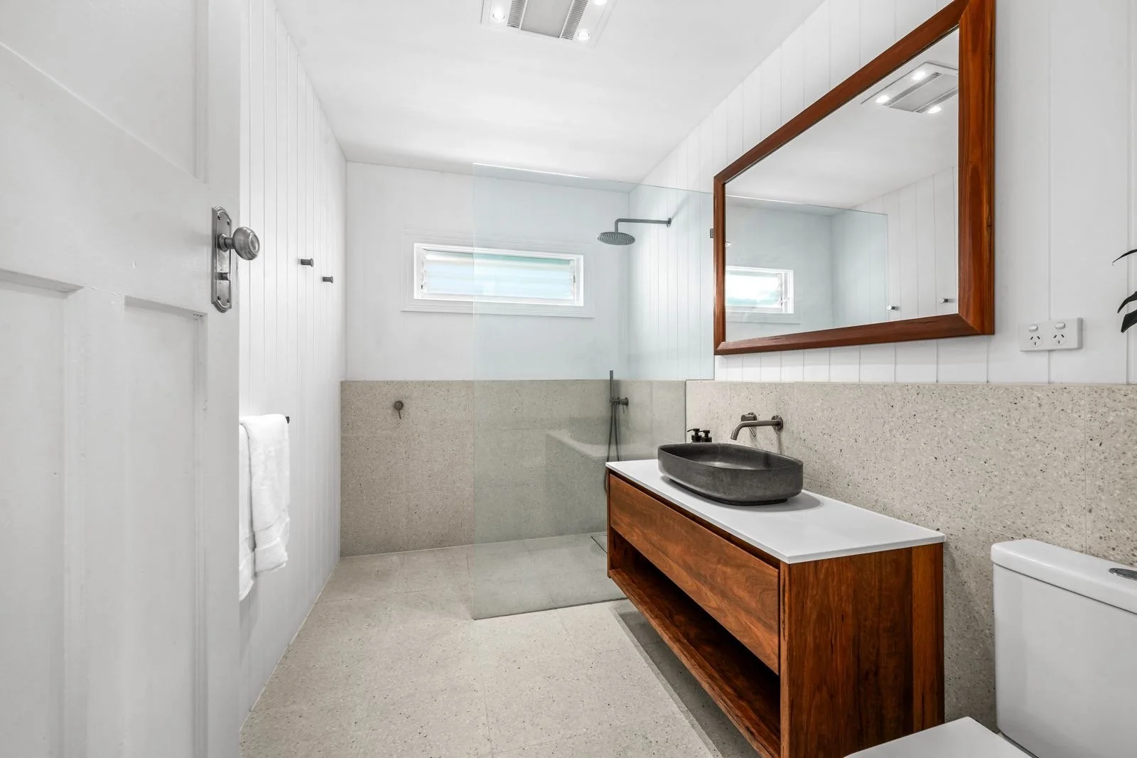 Modern bathroom with white walls, a wooden vanity with a black vessel sink, a large mirror, and a walk-in shower with a glass partition, small rectangular window, and rain showerhead.