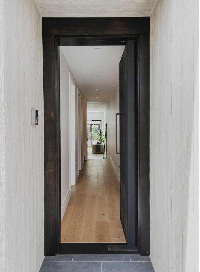 View of a hallway through a partially open black door in a modern home, with light-colored walls, wooden flooring, and a plant at the end of the hall near a window.
