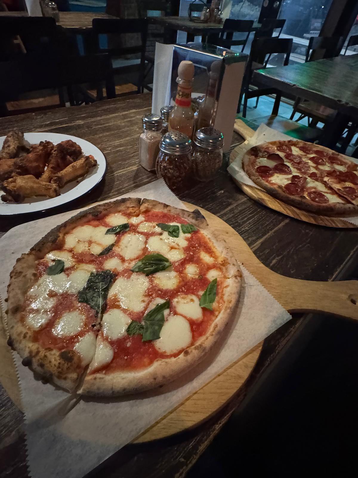 Two pizzas, one with mozzarella, tomato, and basil, and the other with pepperoni, on a wooden table with chicken wings and condiments in the background.