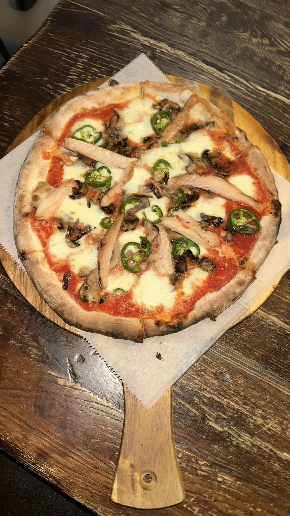 Pizza with cheese, sliced jalapeños, chicken, and mushrooms on a wooden serving board.