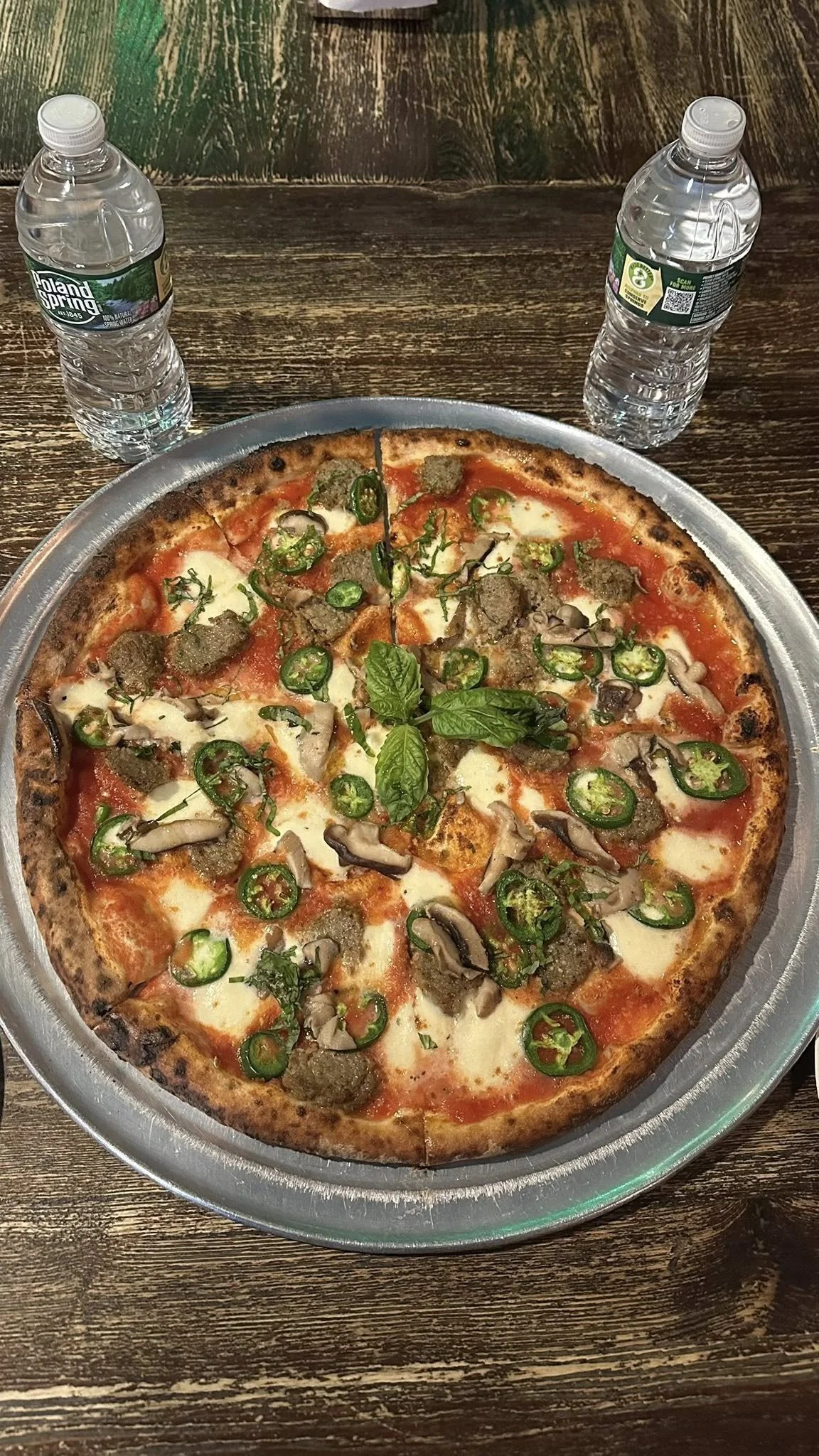 A pizza topped with jalapeños, mushrooms, sausage, and basil, on a round metal tray, with two bottles of water on a wooden table.