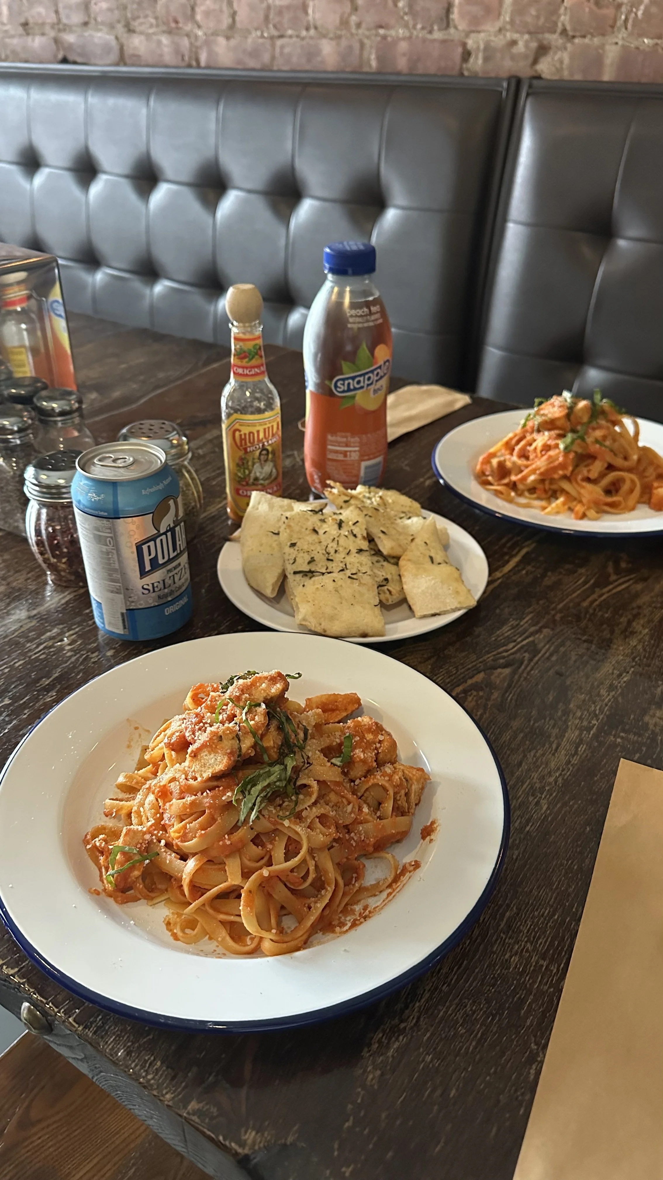 A dining table with two plates of fettuccine Alfredo, a plate of bread with herbs, a can of Polar seltzer, a bottle of hot sauce, a bottle of Snapple peach tea, and various condiments.