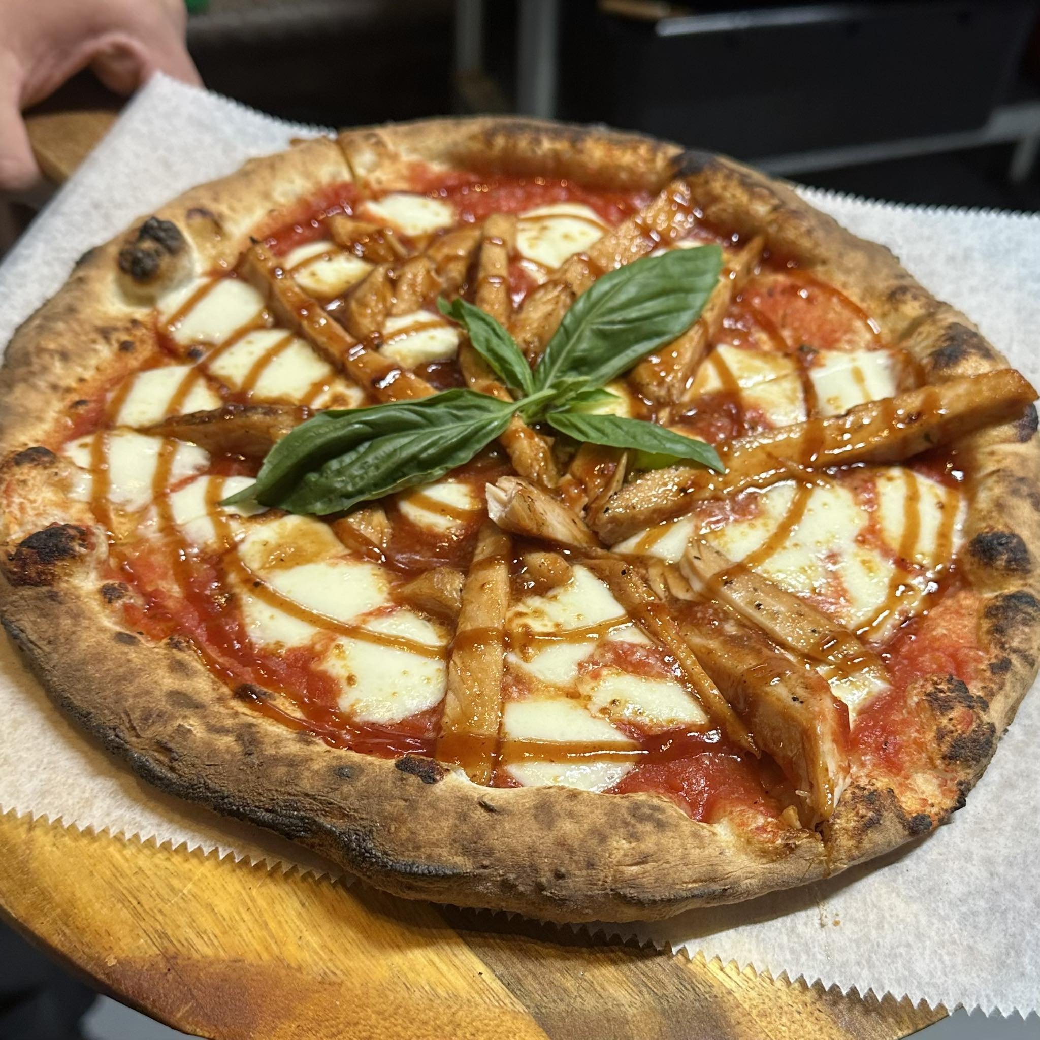 Wood-fired pizza topped with chicken, mozzarella, a basil garnish, and a drizzle of sauce, on a round wooden serving board.