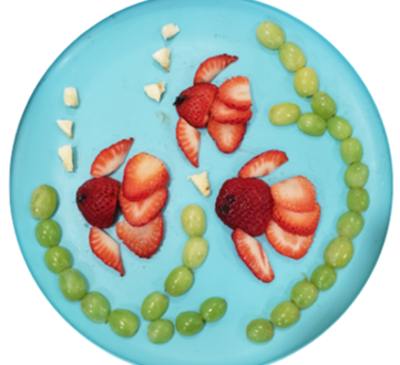 Green grapes, sliced strawberries, and small pieces of pineapple used to make a picture with three fish on a blue plate.