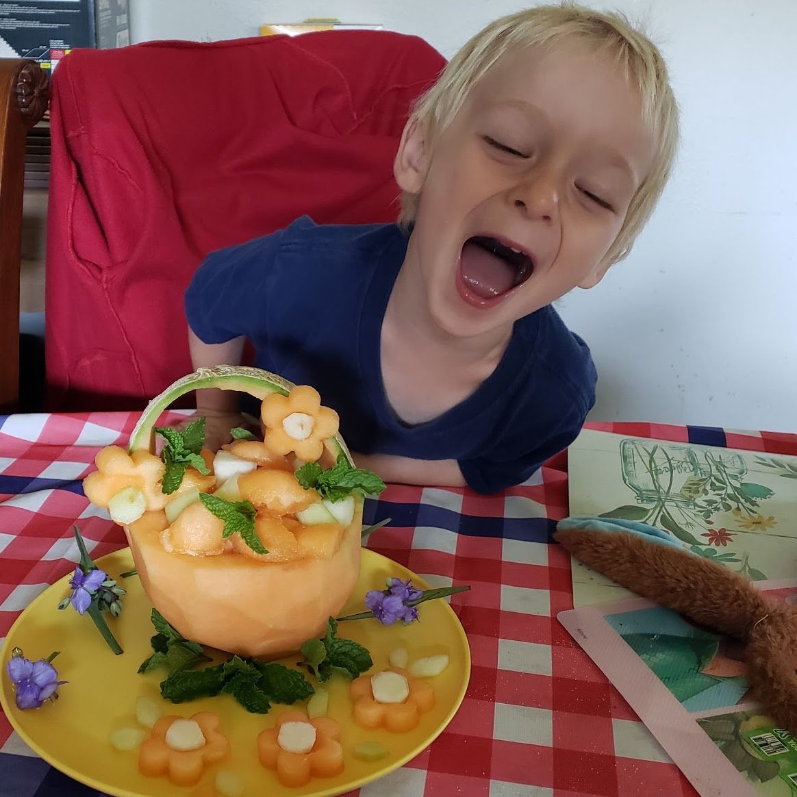 A young boy is excitedly leaning over a colorful fruit platter on a yellow plate. The platter features a melon basket filled with fruits shaped like flowers and mint leaves. The boy is smiling with his mouth wide open, displaying happiness.
