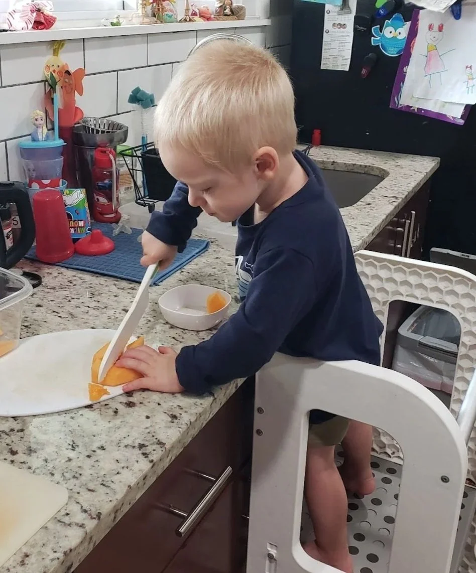 A young boy with blond hair in a navy shirt and shorts is standing in a high chair at a kitchen counter, cutting a piece of cantaloupe with a kid-safe knife.