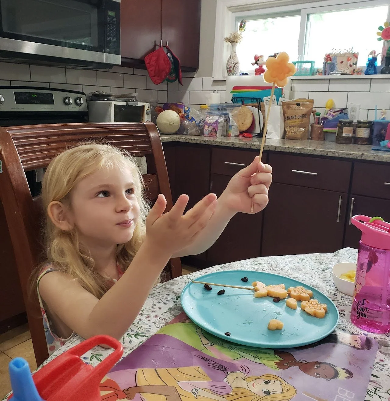 A young girl with blonde hair sitting at a kitchen table, holding a magic wand made of fruit on a stick in the air.