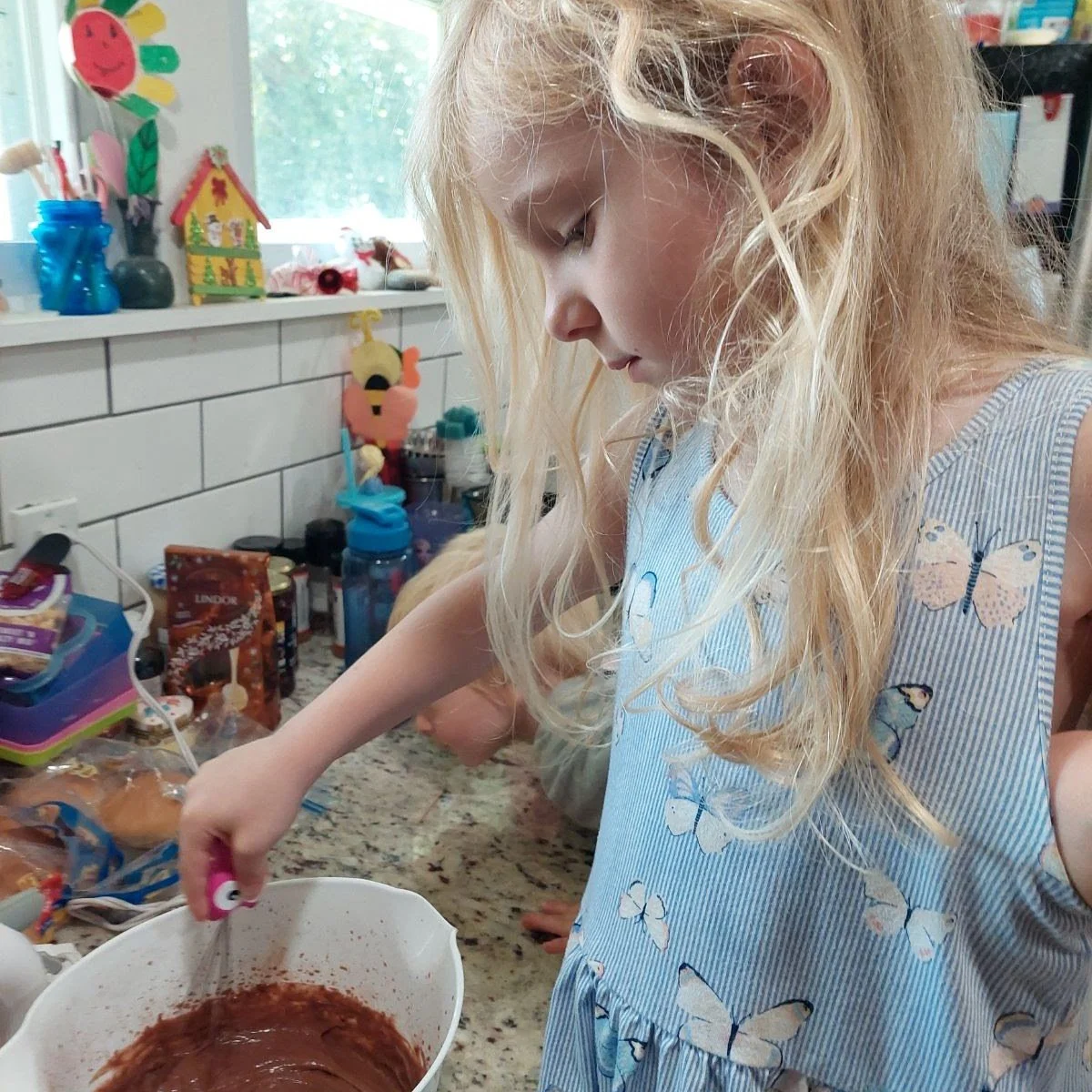 A young girl with long, blonde curly hair wearing a blue striped dress with butterfly prints, mixing food in a white bowl in a kitchen.