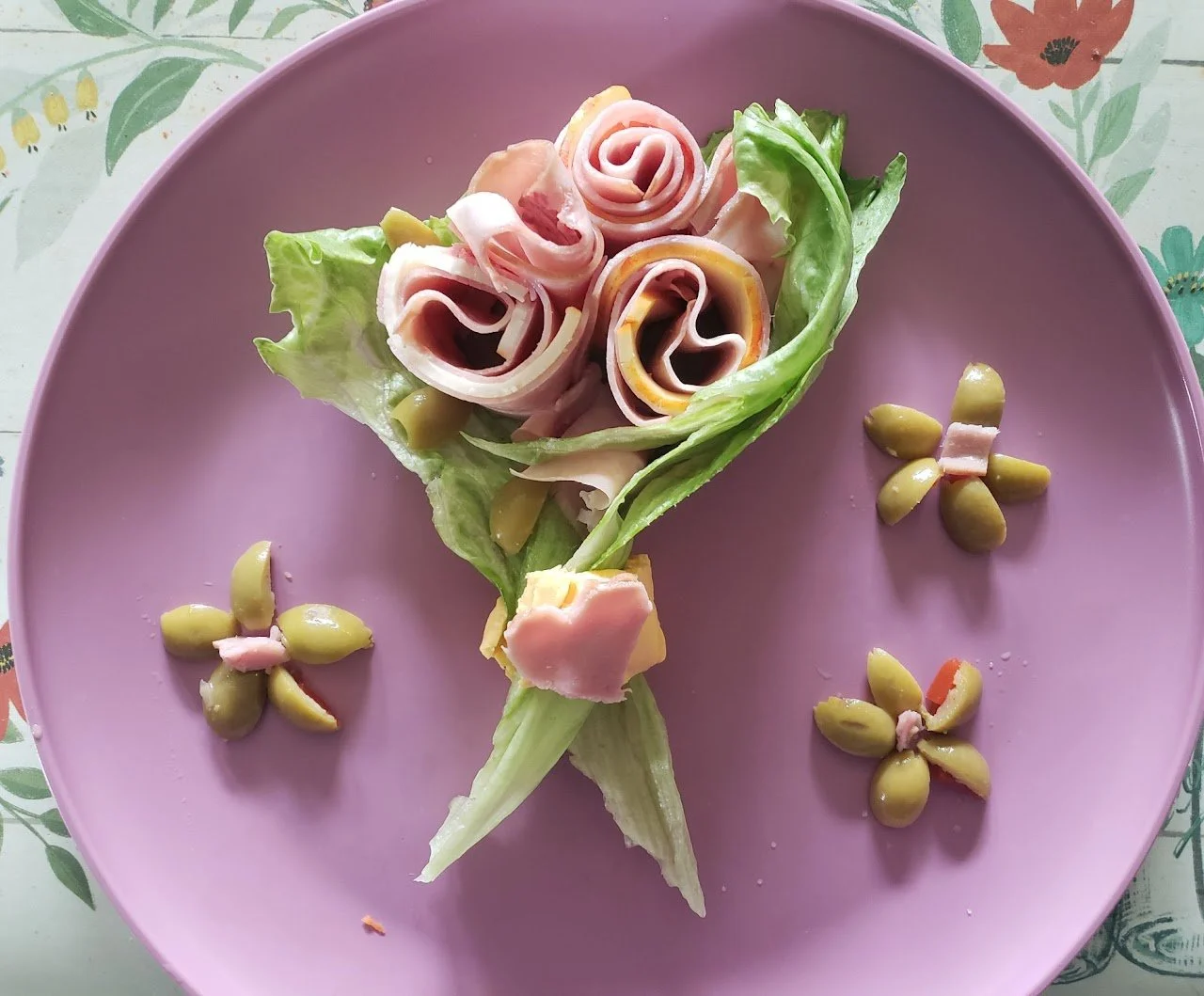 Salad arranged to resemble a bouquet of flowers on a pink plate, with lettuce and rolled slices of ham, and cheese, along with small flower-shaped garnishes made from green olives and ham.