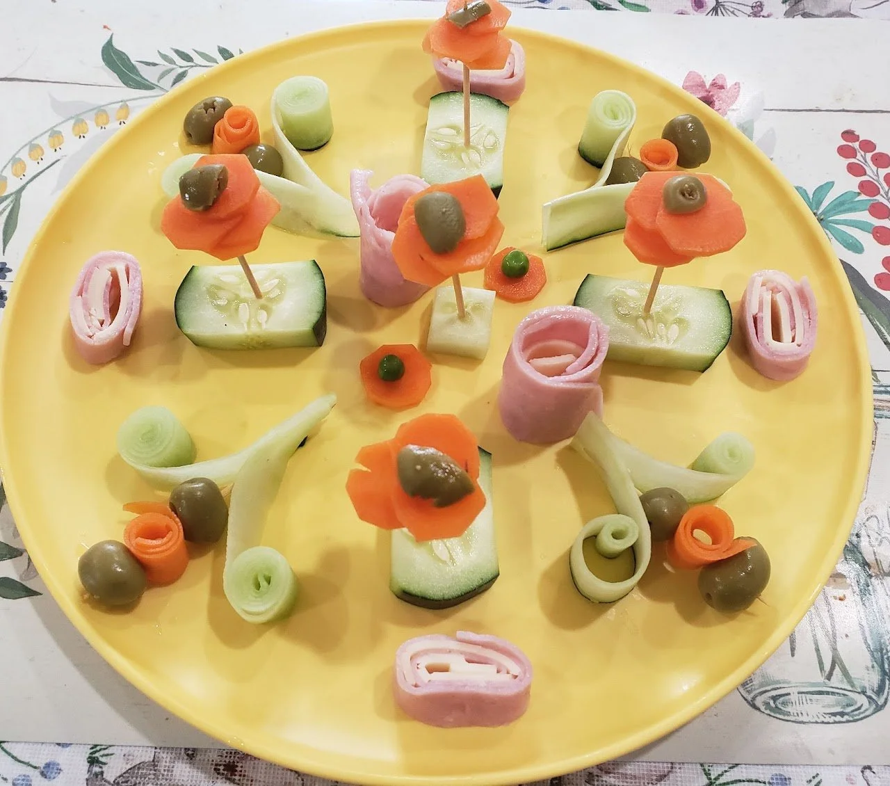 A yellow plate with various vegetables arranged as flowers. Slices of cucumber, rolled ham, carrot slices, and green olives are used to create colorful garnishes.