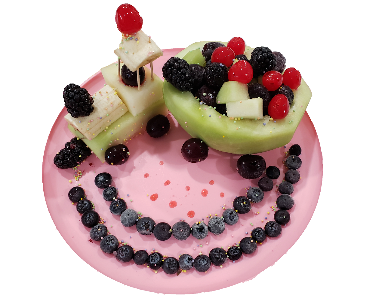 Fruit arrangement that looks like a train on a pink plate, featuring a train made of melon filled with mixed berries and cubed melon pieces, with banana and berry snacks, outlined by a circle of blueberries.