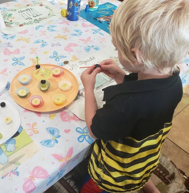 A young boy with blonde hair wearing a black and yellow striped shirt standing at a table, making fruit cupcakes made of grapes, cantaloupe and blueberries.