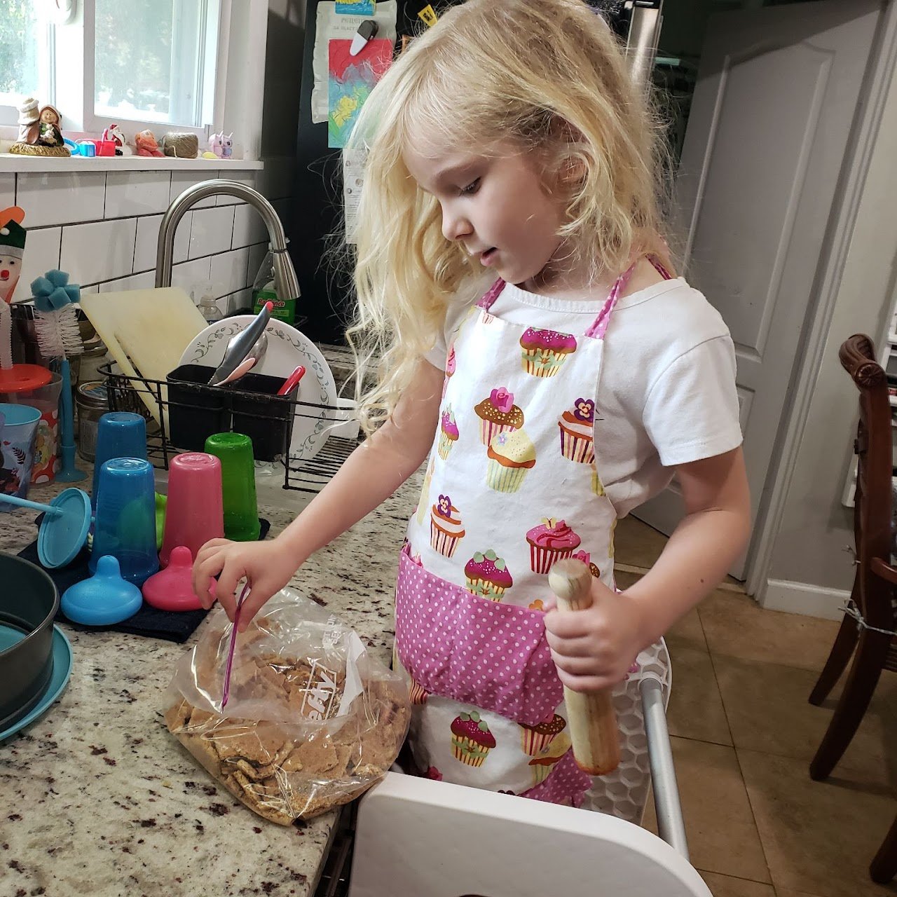 A young girl with blonde hair wearing a cupcake-patterned apron cooking in a kitchen.