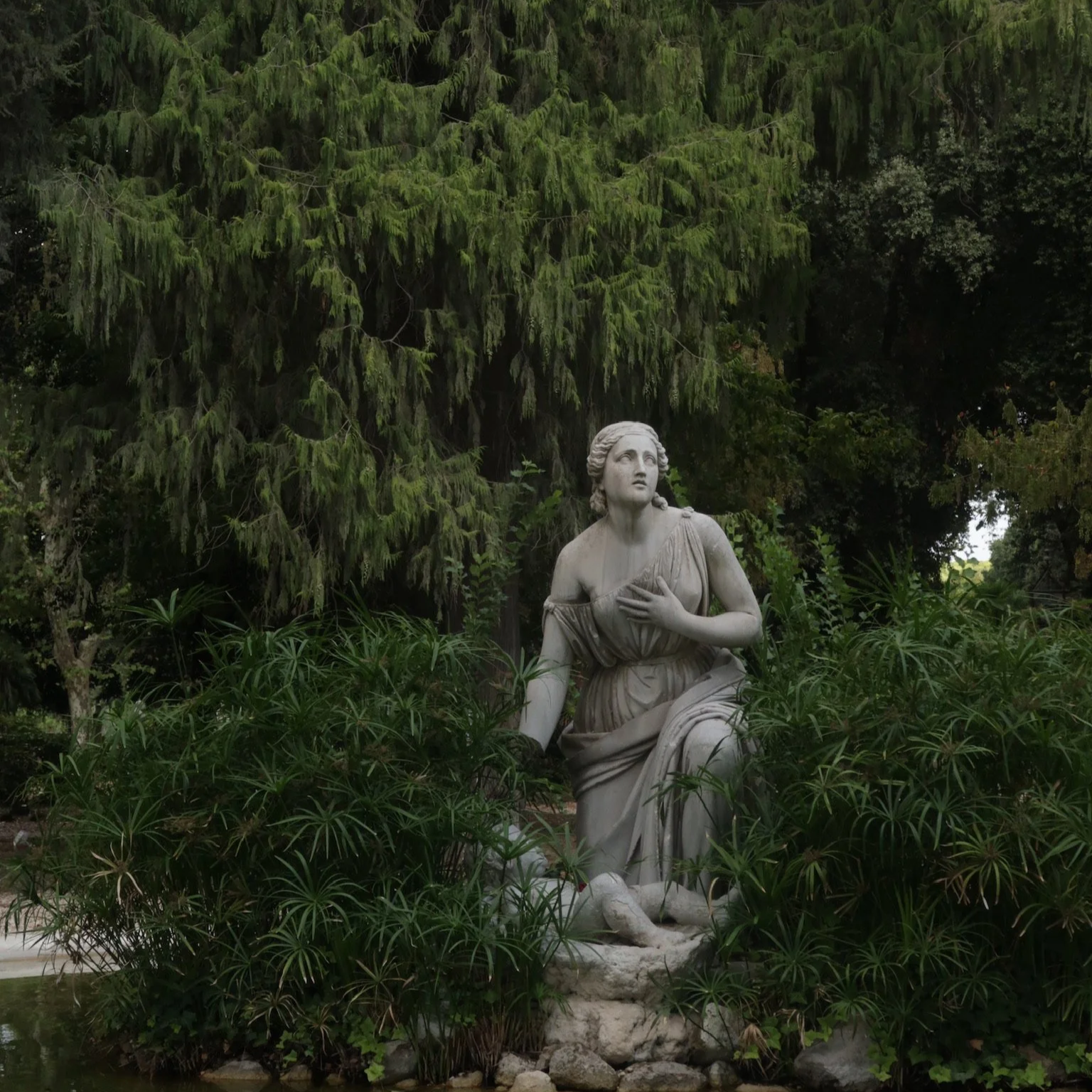 A marble statue of a woman kneeling in a lush garden surrounded by green foliage, with trees in the background.