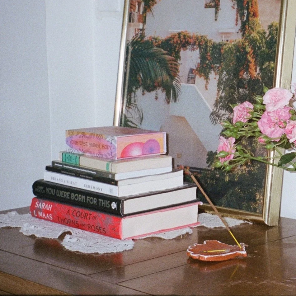 A stack of seven books on a wooden table, with a small flower-shaped incense holder and a stick of incense, a lace doily underneath, and a framed picture of a scenic outdoor view with trees and buildings in the background.