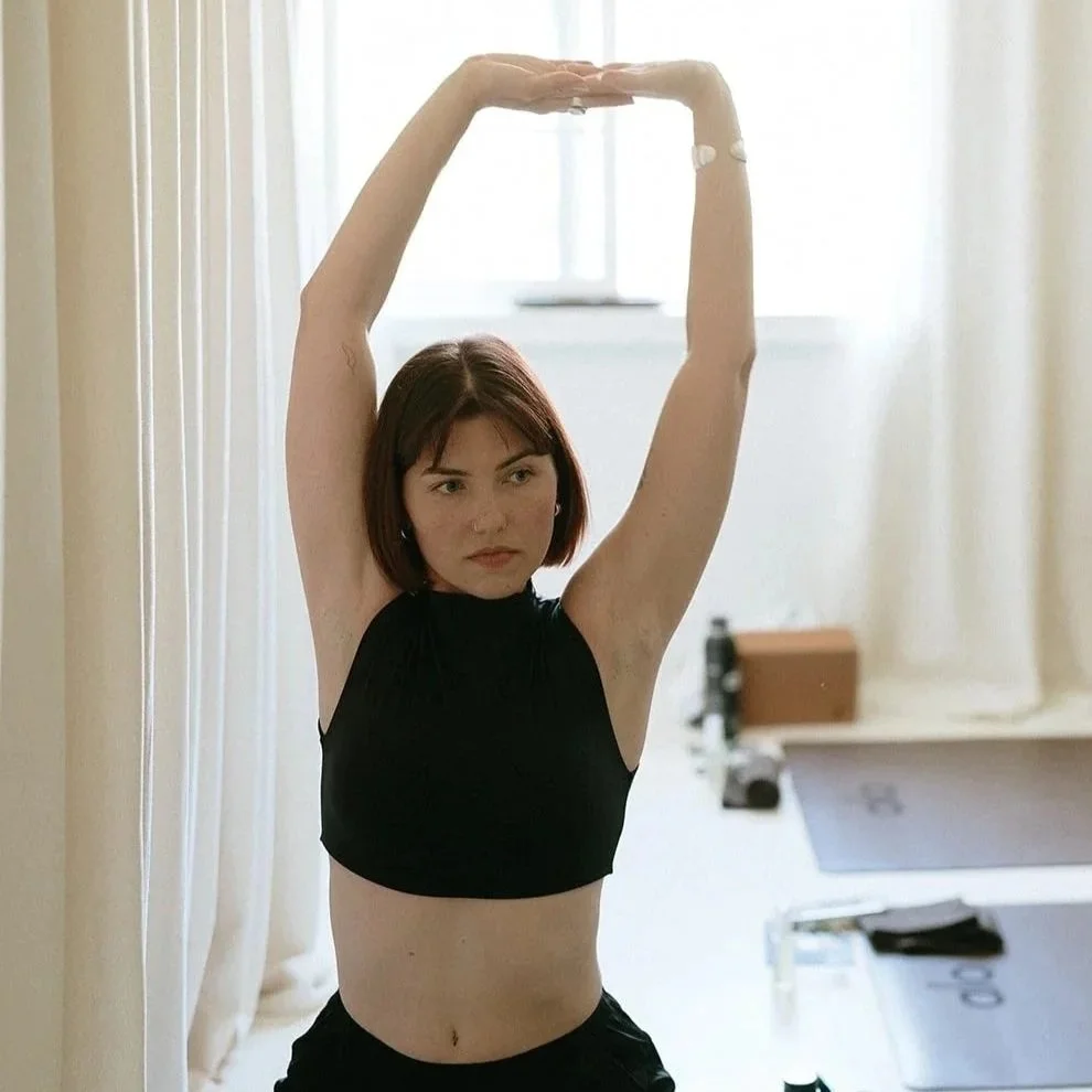 A young woman with short reddish-brown hair practicing yoga in a bright room with sunlight, wearing a black crop top and black pants, standing with arms raised above her head.