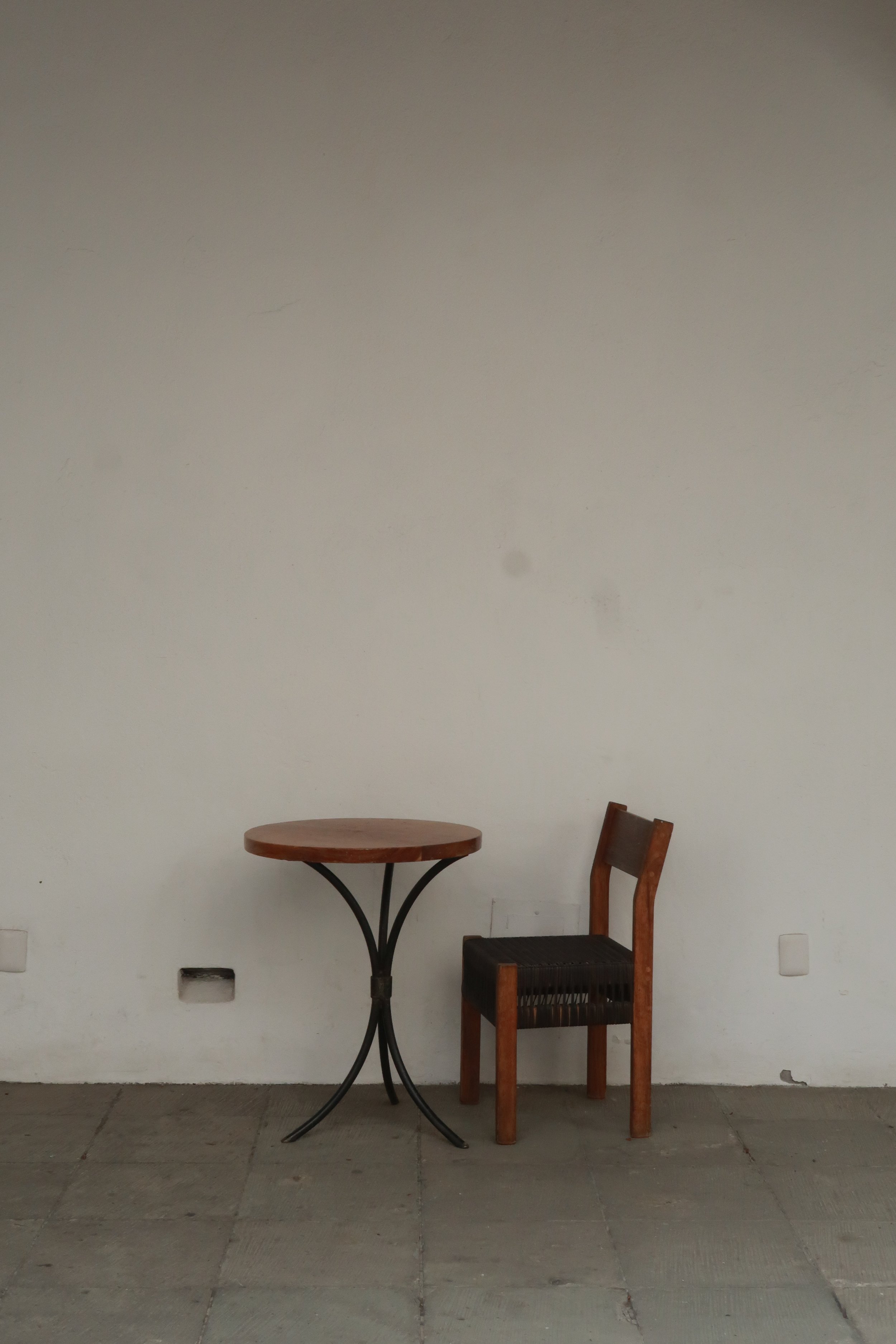 A small round wooden table with a black metal base next to a wooden chair with a woven seat, against a plain white wall on a tiled floor.