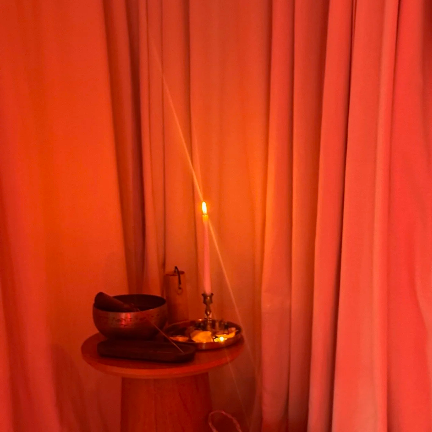 A small altar with a lit candle, a singing bowl, and a small wooden object on a wooden table with closed red curtains in the background.