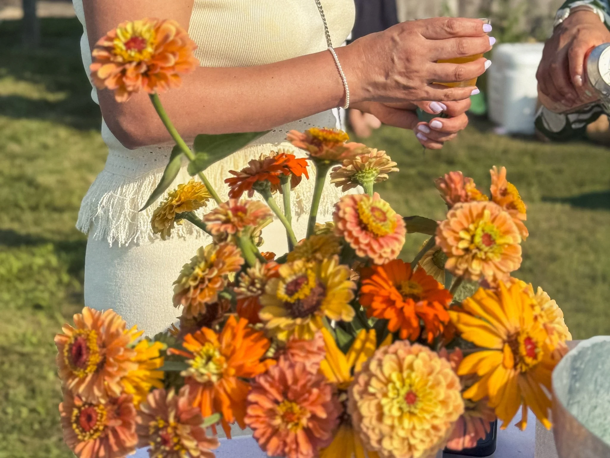 Person holding a glass of beer standing outdoors near a bouquet of orange, yellow, and pink flowers.